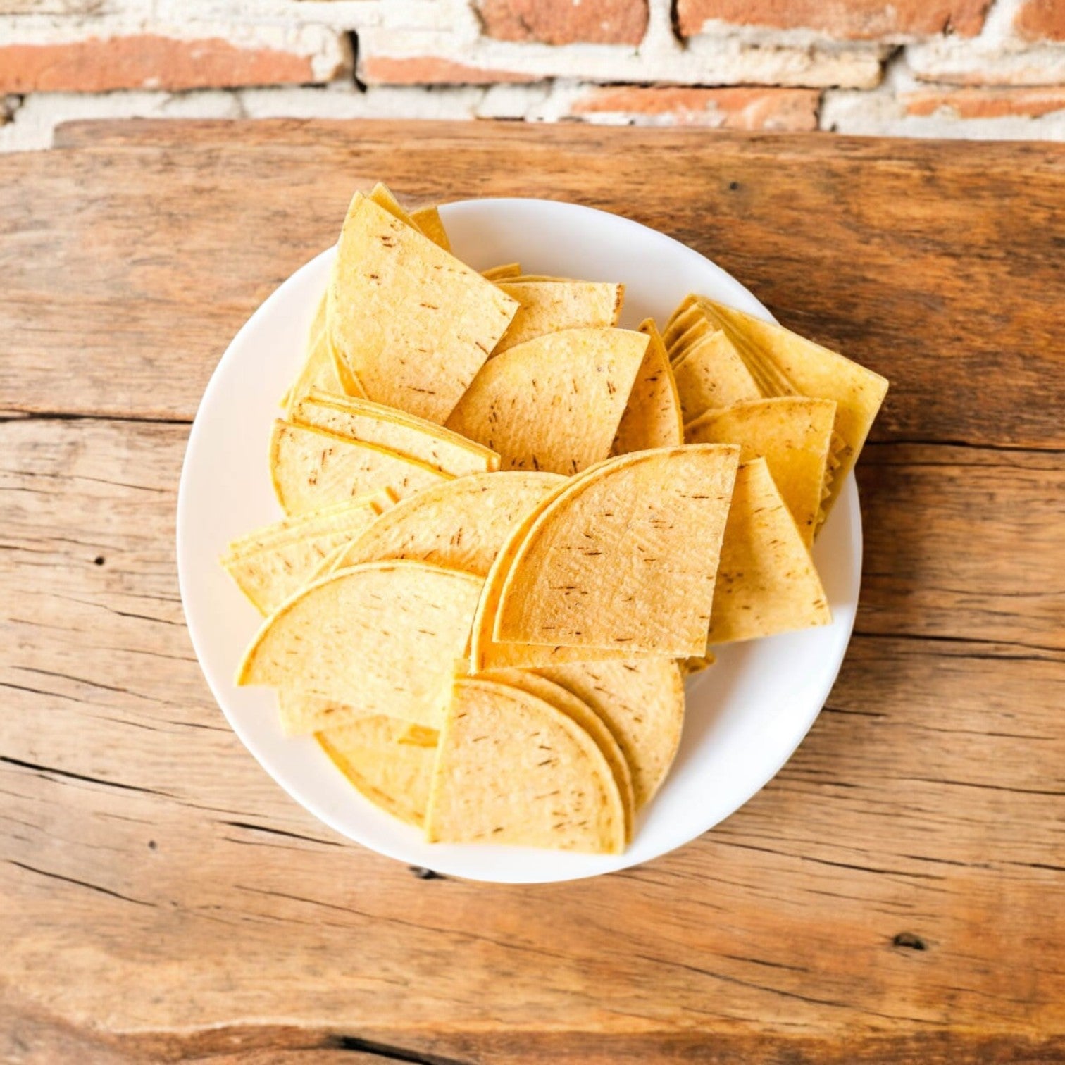 A rustic wooden table displays a white plate of neatly arranged, halved El Milagro White Corn Tortillas (4-Way Cut, 20 lbs). Note: Ships Mondays. Plan accordingly! Brick wall in the background.