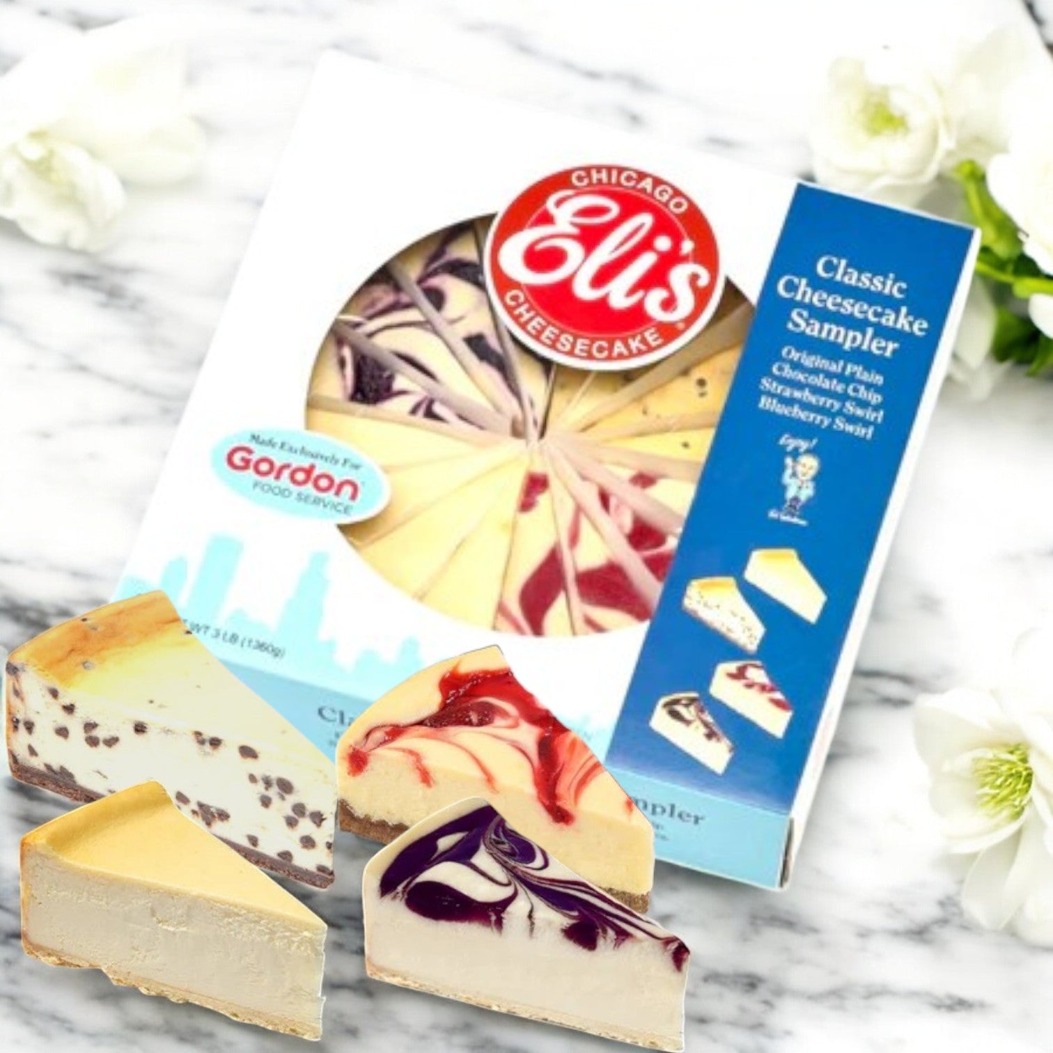 A box of Eli's Cheesecake 9" Classic Cheesecake Sampler, offering assorted classic cheesecake slices, sits on a marble surface adorned with white flowers.