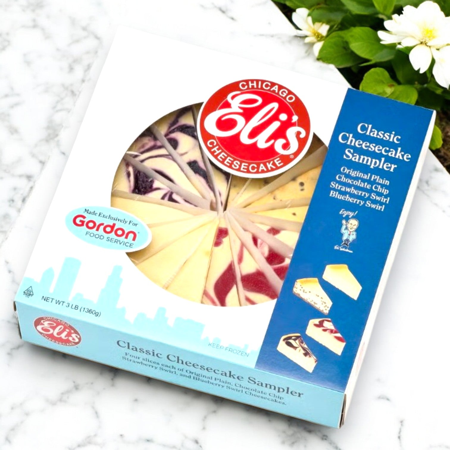 A box of Eli's Cheesecake's 9" Classic Cheesecake Sampler - 3 Count sits on a white surface, showcasing plain, chocolate chip, strawberry swirl, and blueberry swirl slices.