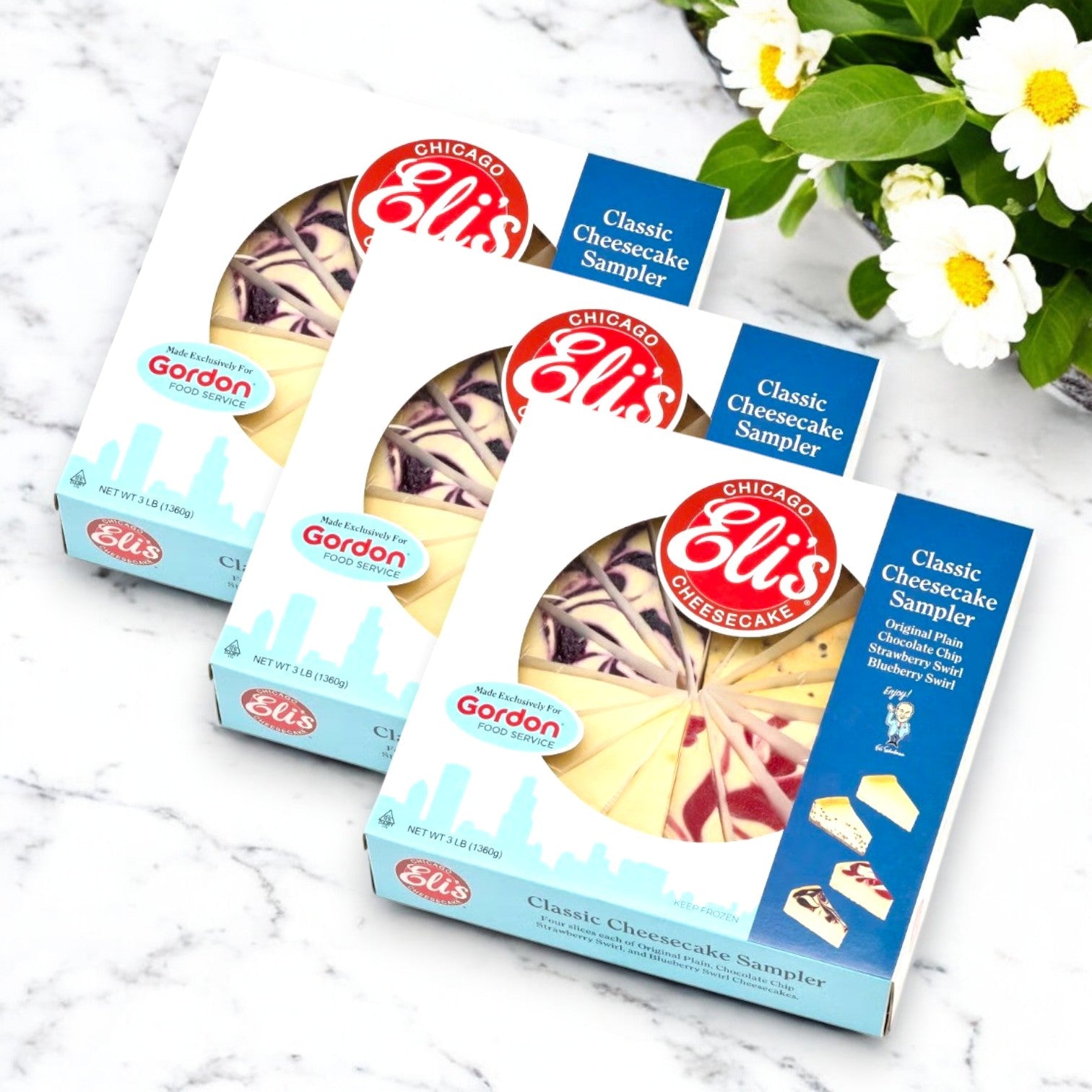 Three boxes of Eli's Cheesecake 9" Classic Cheesecake Sampler, each featuring assorted slices in blue packaging, are on display with white flowers and green leaves enhancing the scene.
