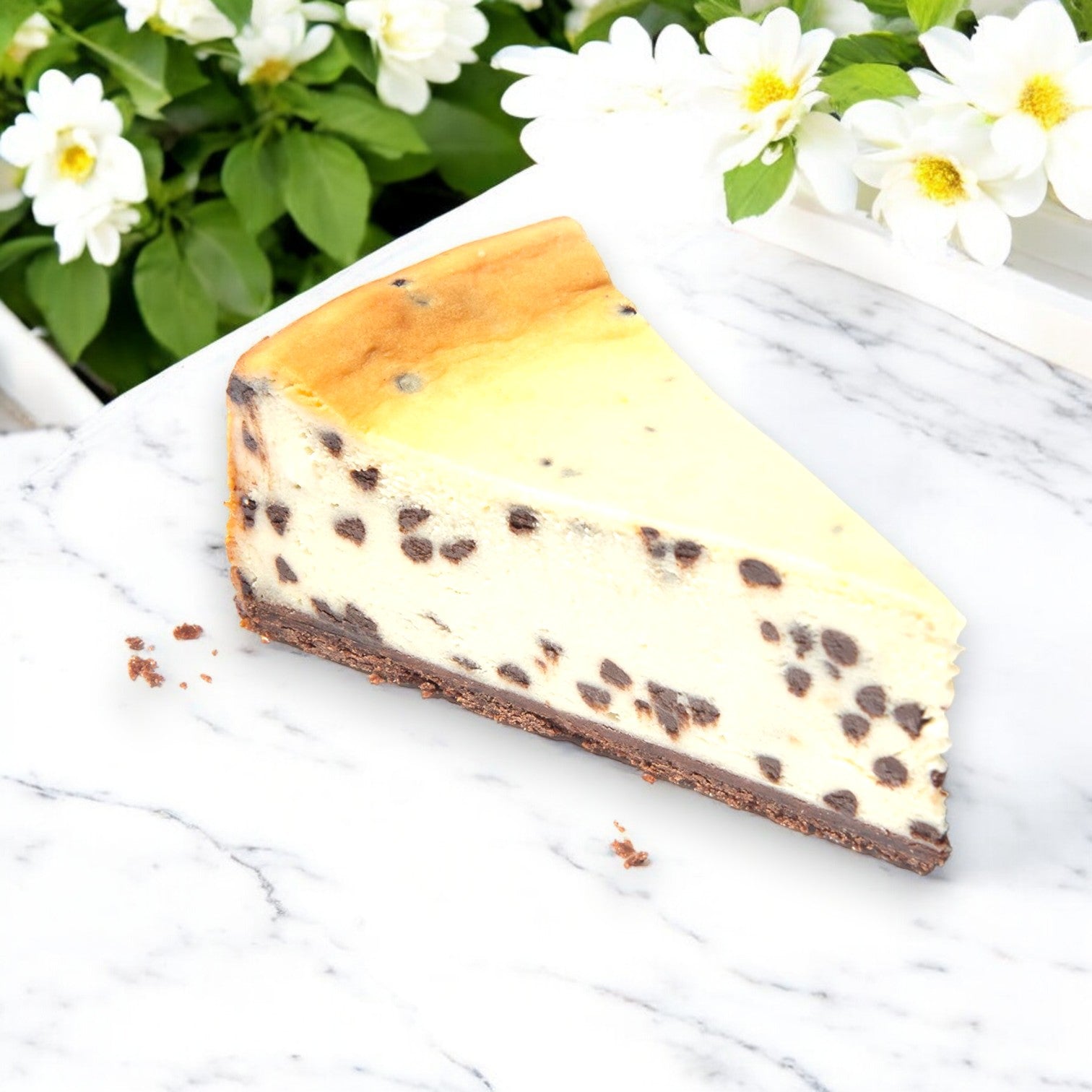 A slice of Eli's Cheesecake 9" Classic Sampler elegantly sits on a marble background adorned with delicate white flowers.