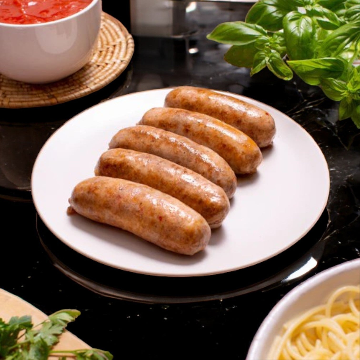 Five of Esposito's Hot Italian Sausages sit on a white plate, next to a bowl of red sauce, basil, parsley, and cooked spaghetti on a dark countertop. Enjoy authentic Italian flavor with every bite made from premium ingredients.