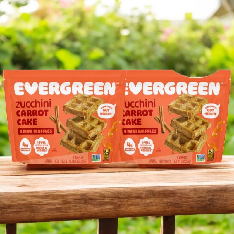 Two packages of Evergreen Zucchini Carrot Cake Frozen Mini Waffles (9 oz., 2 count) are shown outdoors on a wooden surface with greenery, showcasing this delicious and nutritious breakfast option from Evergreen.