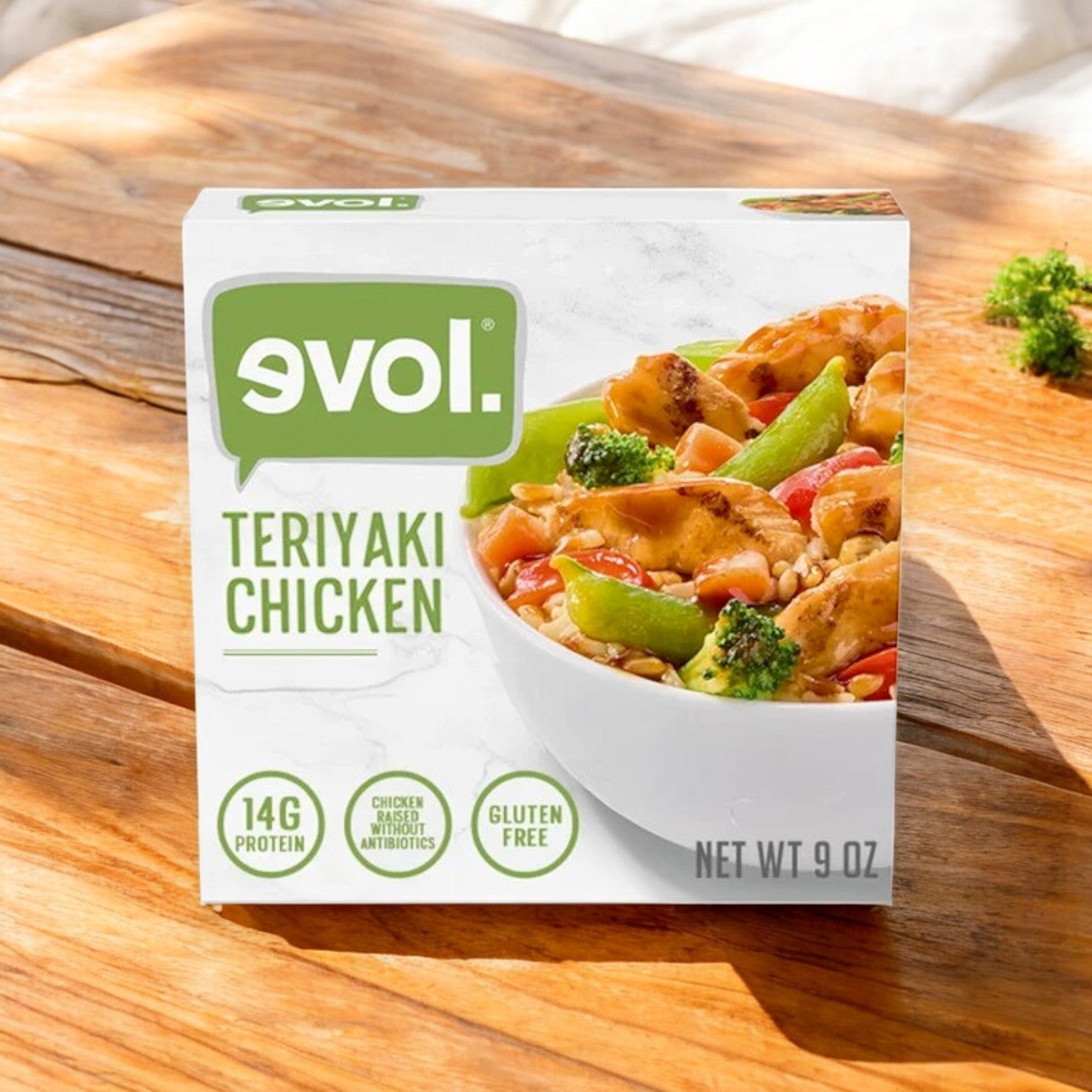 A 9 oz Evol Teriyaki Chicken Frozen Meal sits on a wooden surface, featuring chicken, broccoli, red bell peppers, and rice. It boasts 14g protein and a gluten-free label, alongside varieties like butternut squash and vegetarian ravioli for diverse meal choices.