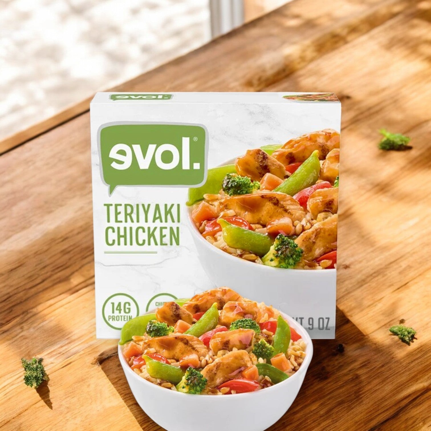 A 9 oz Evol Teriyaki Chicken Frozen Meal rests on a wooden surface, while a bowl showcases its colorful contents: chicken, bell peppers, and broccoli. This vibrant medley pairs perfectly with butternut squash or sage ravioli.