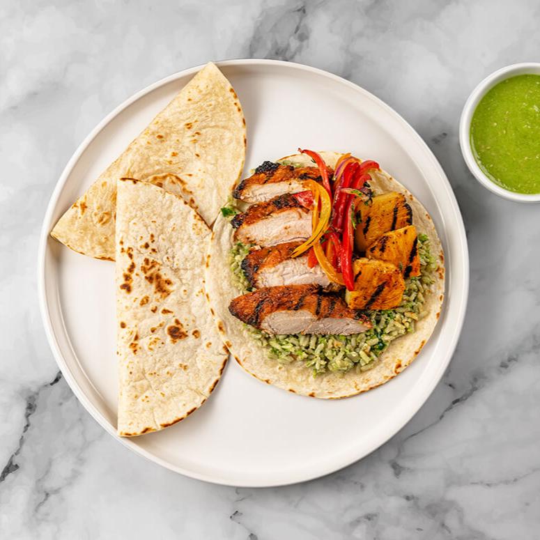 A single serving of FoodHaul Al Pastor Chicken Tacos, featuring grilled chicken, rice, and grilled vegetables encased in tortillas with a side of green sauce, captures authentic Mexican flavors on a marble surface.
