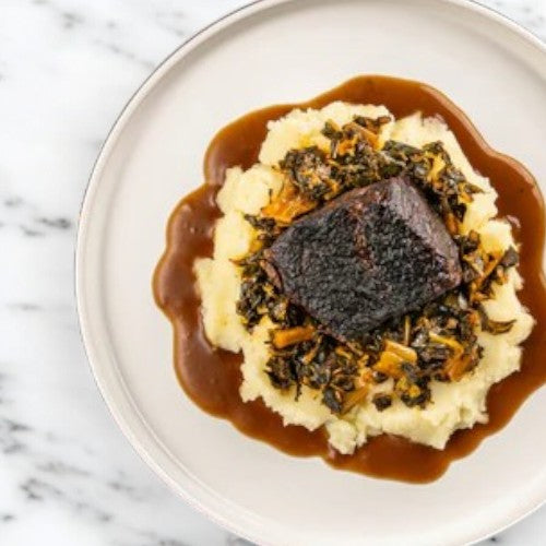 Indulge in Easy Lunches' FoodHaul Braised Short Ribs, featuring tender short ribs and flavorful braised greens on a bed of creamy mashed potatoes, all enhanced by a rich brown sauce.