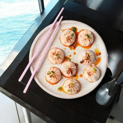 The Mila 3 Pack Soup Dumpling Combo, rich with authentic flavors and herbs, is set on a dark table by a window. Pink chopsticks and a spoon lie beside it, tempting you to enjoy every bite.