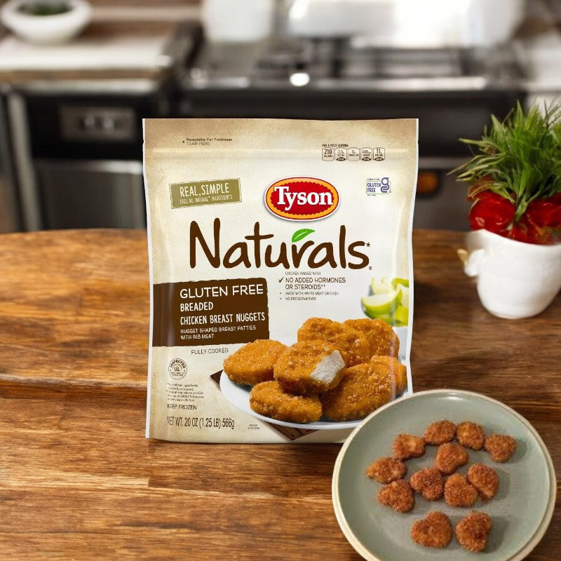 A 20 oz. bag of Tyson Naturals Gluten Free Breaded Chicken Breast Nuggets is on a wooden table with a plate of crispy nuggets. The slightly blurred kitchen background highlights this delicious Tyson meal.