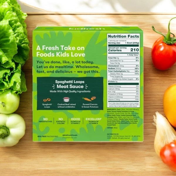 A box of KidFresh Frozen Spaghetti Loops With Meat Sauce, 7.25 oz. -3 Count, rests on a wooden surface amidst various vegetables. The packaging emphasizes its nutritious ingredients like hidden veggies and showcases a detailed nutrition facts label.