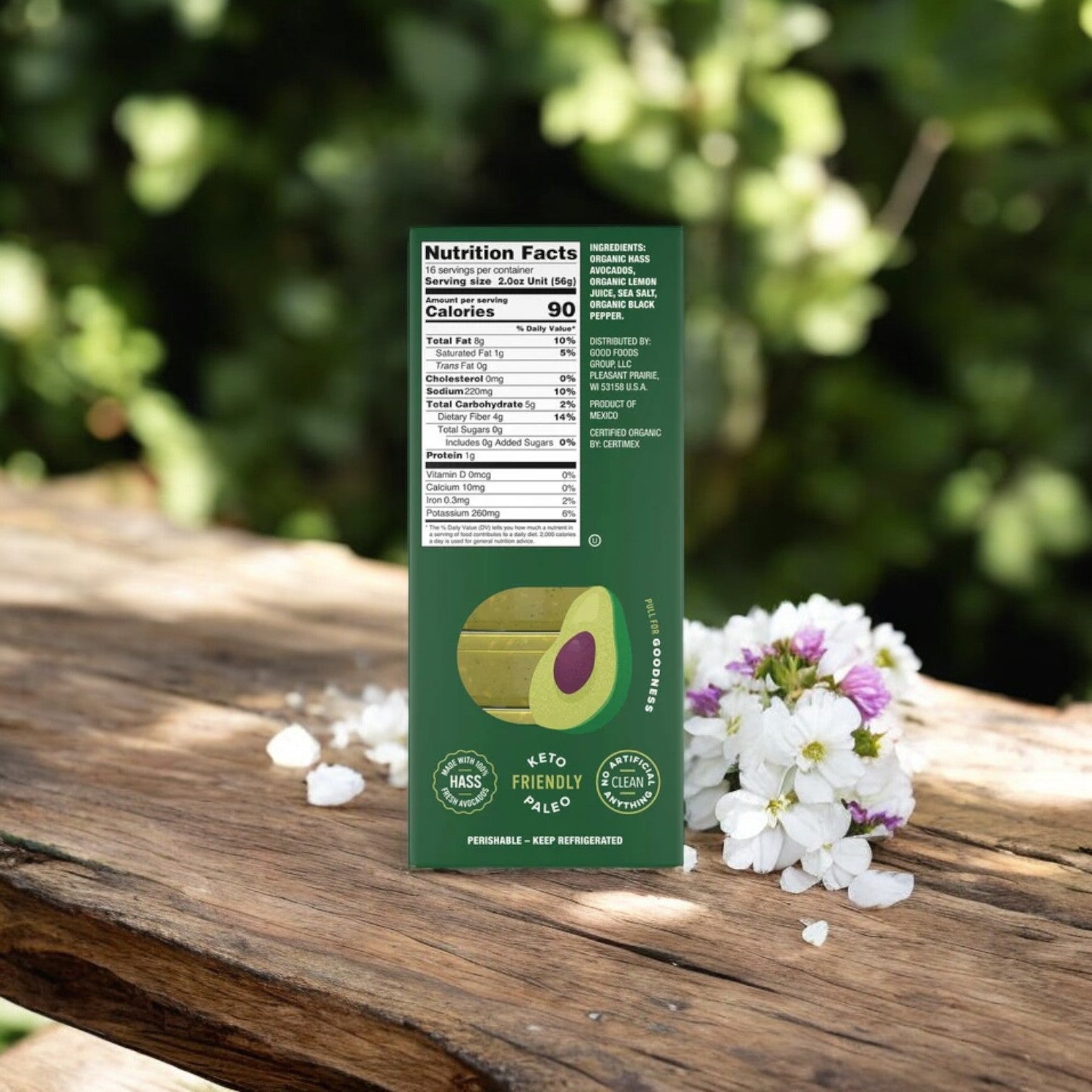 A carton of Good Foods Organic Avocado Mash, Sea Salt & Black Pepper, rests on a wooden surface, surrounded by white and purple flowers. The packaging proudly displays its nutrition facts and labels, featuring "Keto Friendly," "Non-GMO," and "Gluten-Free.