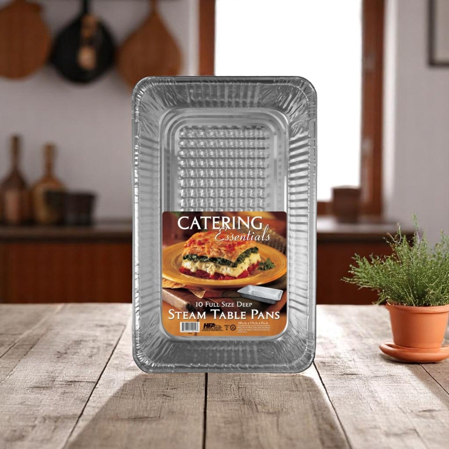 A 10-pack of Handi-Foil of America HFA Deep Full-Size Steam Table Pans sits on a kitchen counter, with a plant nearby and utensils hanging in the background.