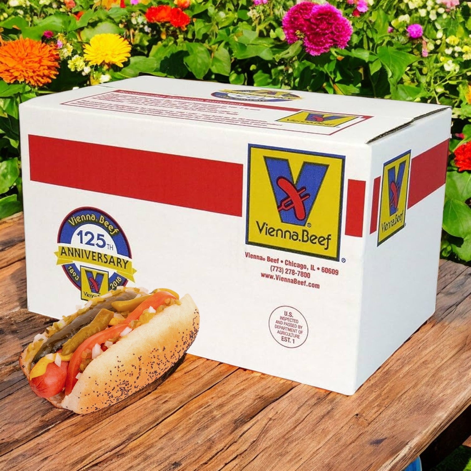 A Vienna Beef All-Beef 4:1 Hot Dog with smoked flavor and toppings rests on a wooden surface before a branded box, surrounded by colorful flowers.