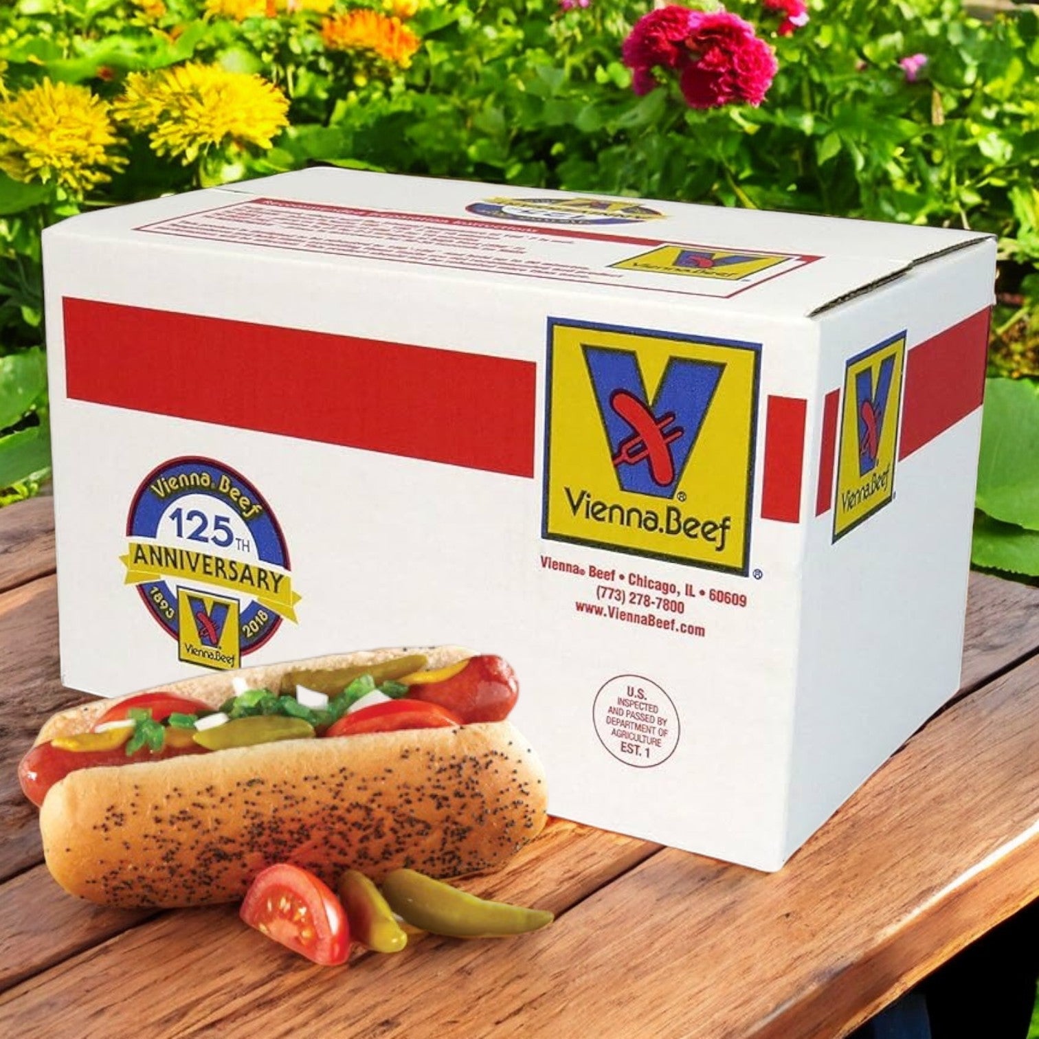 A box of Vienna Beef All-Beef 5:1 6" Skinless Smoked Cooked Hot Dogs rests near a smoked hot dog on an outdoor table surrounded by blooming flowers, proudly displaying the "125th Anniversary" logo and Vienna Beef details.