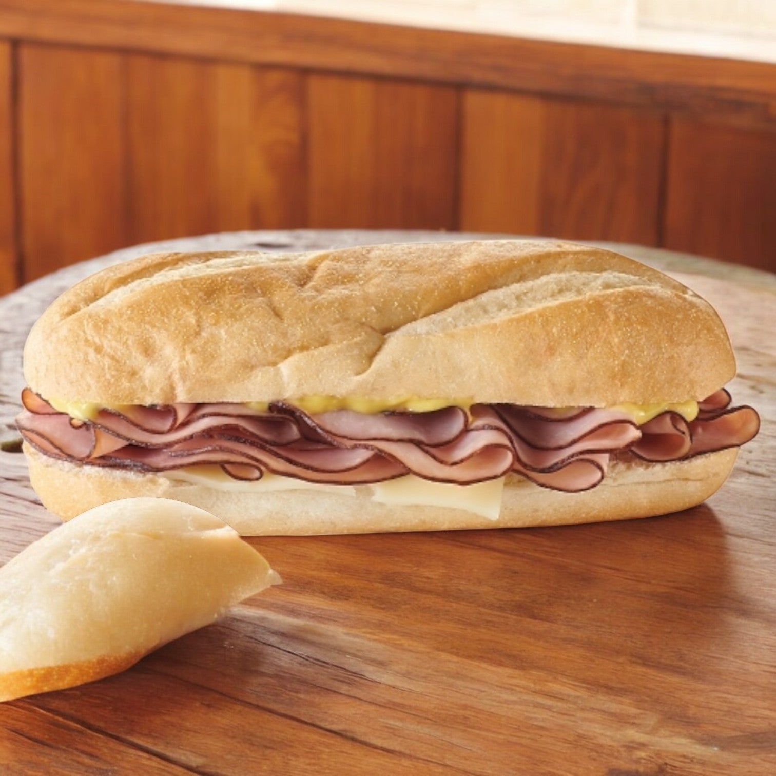 An E.A. Sween Smoked Ham & Swiss Cheese Sub, known for its 30-day shelf life after defrosting, sits on a wooden table with mustard and a crusty baguette finish.