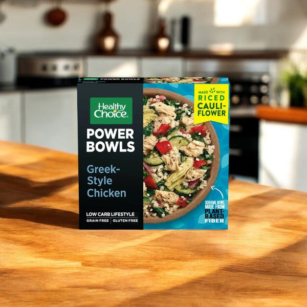 A Healthy Choice Power Bowls Variety Pack sits on a wooden kitchen counter, highlighting the microwaveable convenience of its Greek-Style Chicken flavor with nutritious riced cauliflower, low-carb, and gluten-free features.