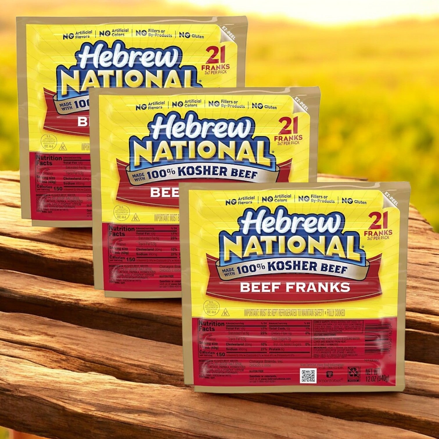 Displayed on a wooden surface with a field in the background, three packs of Hebrew National Beef Franks 12 oz., 3 ct. - 3 Pack promise a premium hot dog experience.