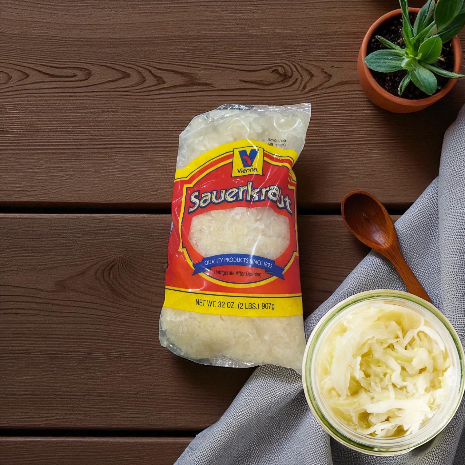 A sealed 32 oz Vienna Beef Sauerkraut bag sits on a wooden table beside a jar of Chicago-style sauerkraut, a wooden spoon, gray cloth, and a potted plant.