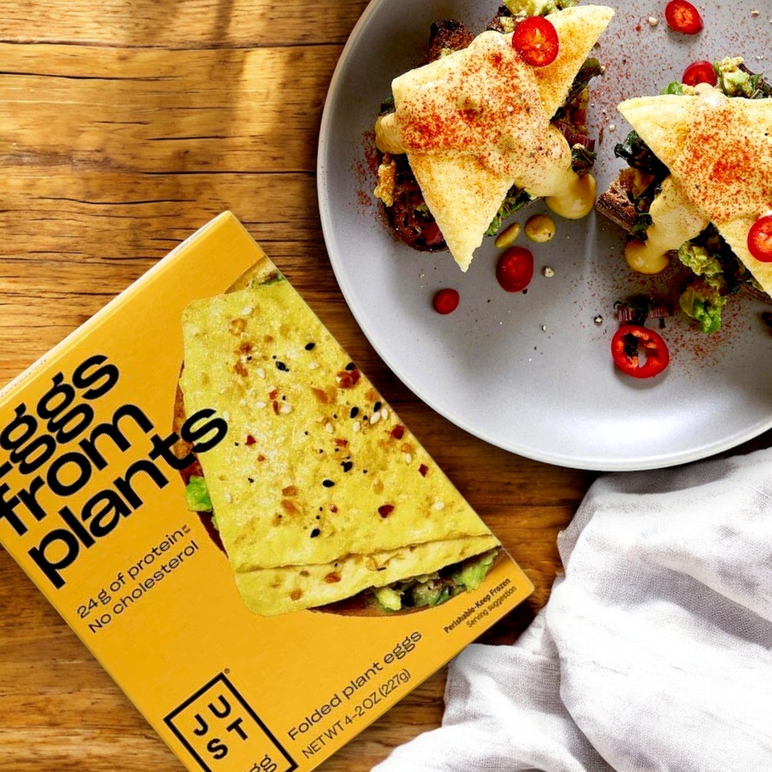 A plate holds two vegan sandwiches topped with cholesterol-free, plant-based eggs. Beside them is a yellow box labeled "Just Egg Folded, plant-based egg, 8 oz. - 1 Pack," made by Just Egg from mung bean protein for a delicious alternative.