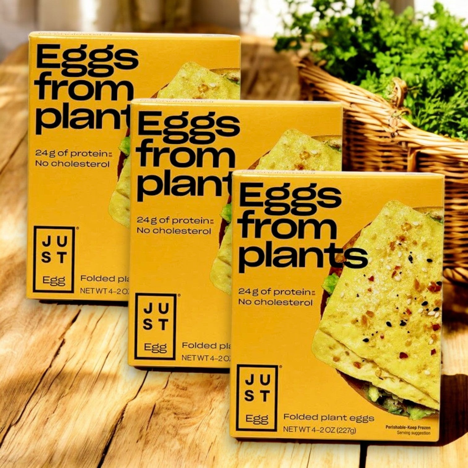 Three boxes of Just Egg Folded, plant-based egg, by Just Egg sit on a wooden table. Made with mung bean protein, they offer 24g of protein and are cholesterol-free. A wicker basket with a green plant decorates the backdrop.
