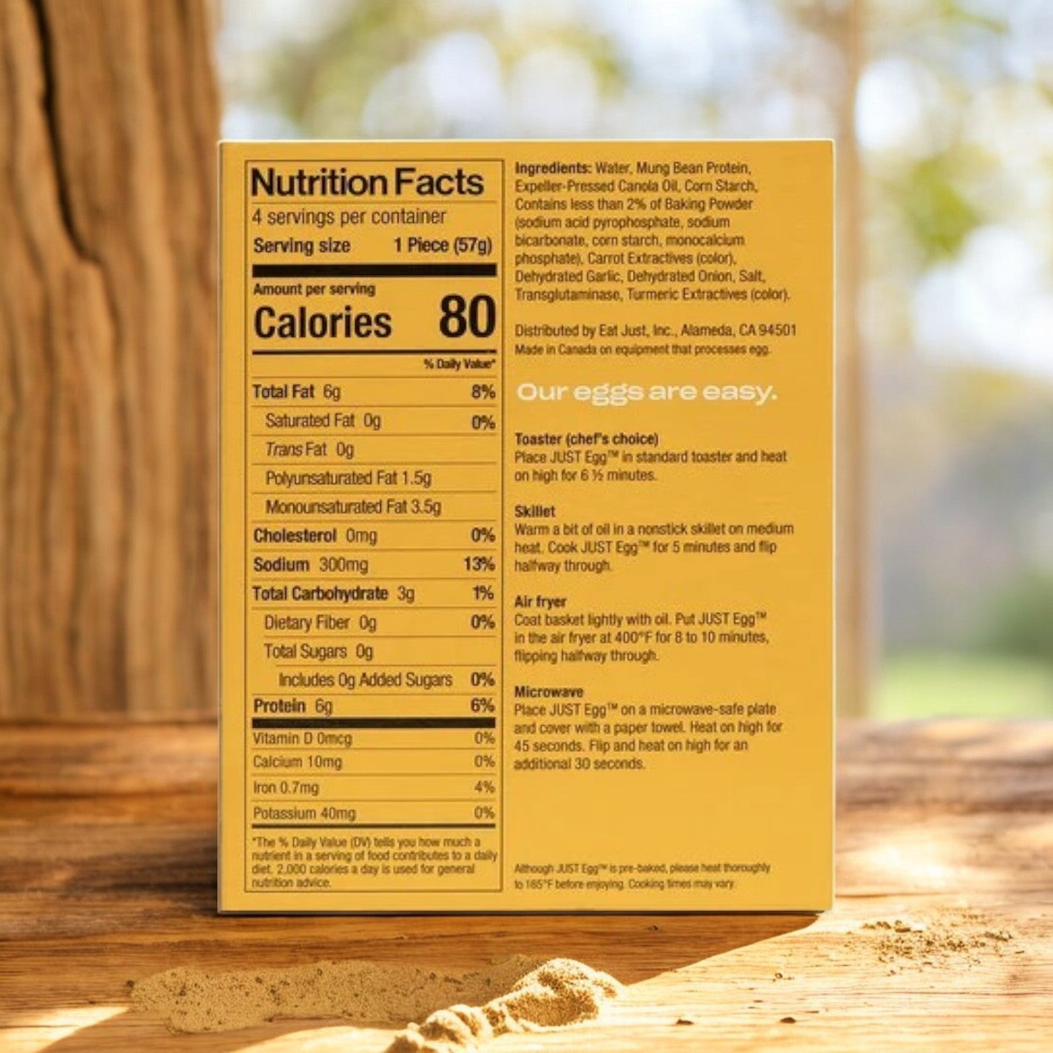 A box of Just Egg Folded, a plant-based egg substitute by Just Egg, sits outdoors on wood. The package highlights its mung bean protein content and includes nutrition facts and cooking instructions against a blurred leafy backdrop.