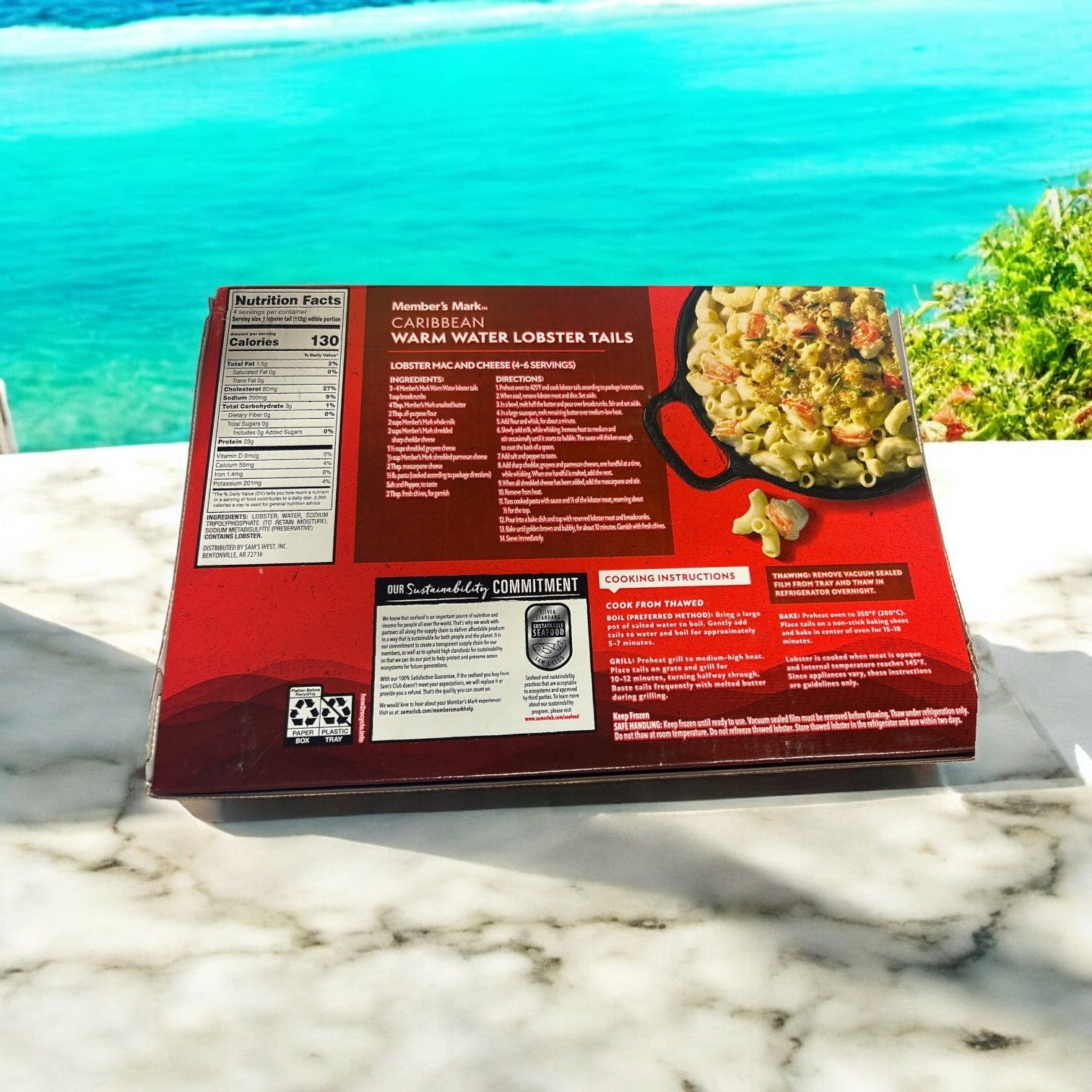 A box of Member's Mark Warm Water Lobster Tails, Frozen (4 Count-3 Pack), sits on a marble table with the blurred ocean and lush foliage in the background.