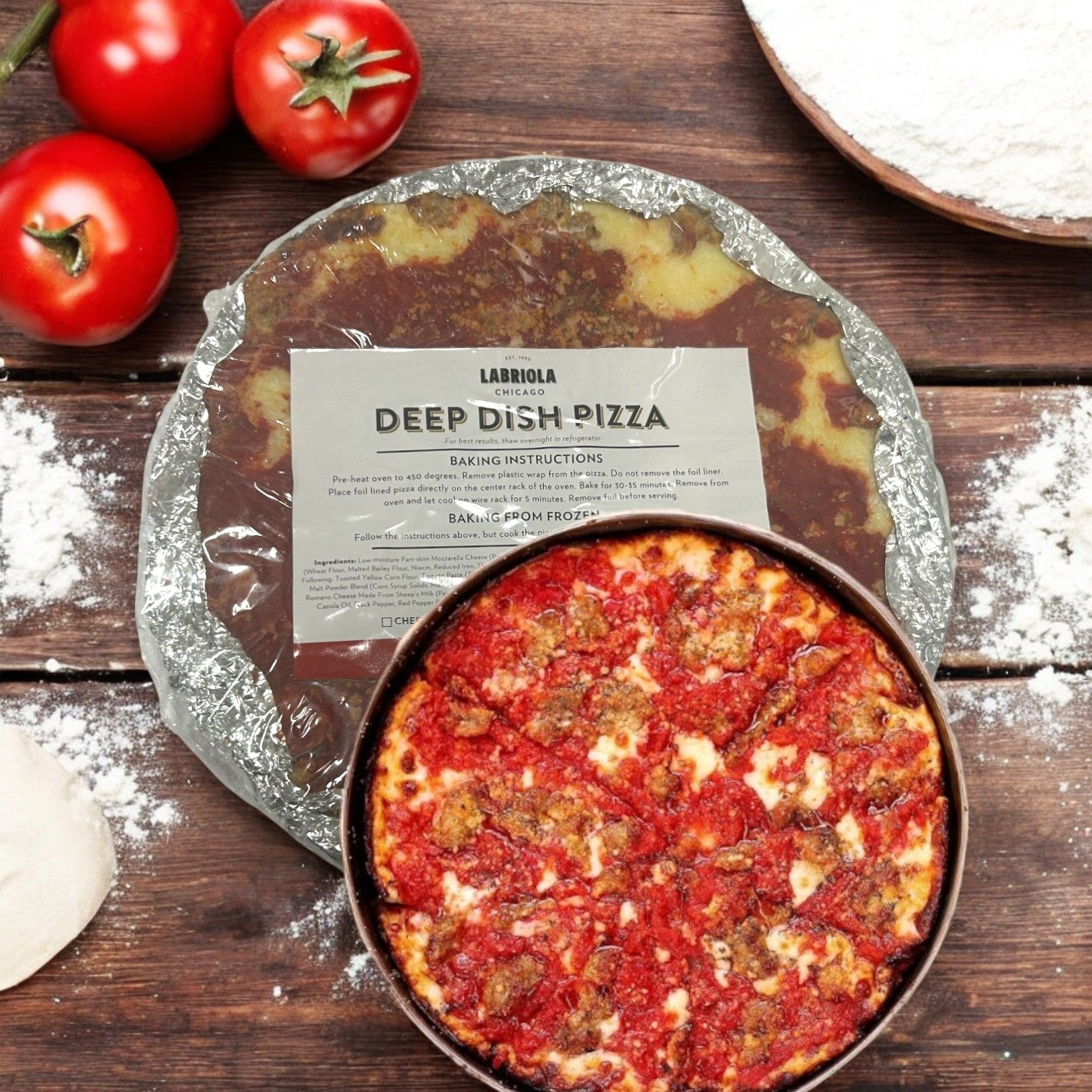 A Labriola Chicago Italian Sausage Deep Dish Pizza-12Inch-1 Count sits next to an unbaked pizza, with fresh tomatoes and a bowl of flour on a wooden surface. Its buttery crust and gooey mozzarella cheese promise a delectable feast.