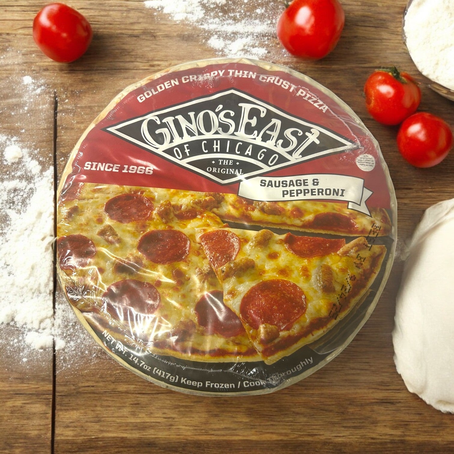 A 3-pack of Gino's East of Chicago Sausage & Pepperoni Deep Dish Pizza (14.7 oz each, total 50.1 oz) is placed on a wooden surface surrounded by tomatoes, flour, and dough.