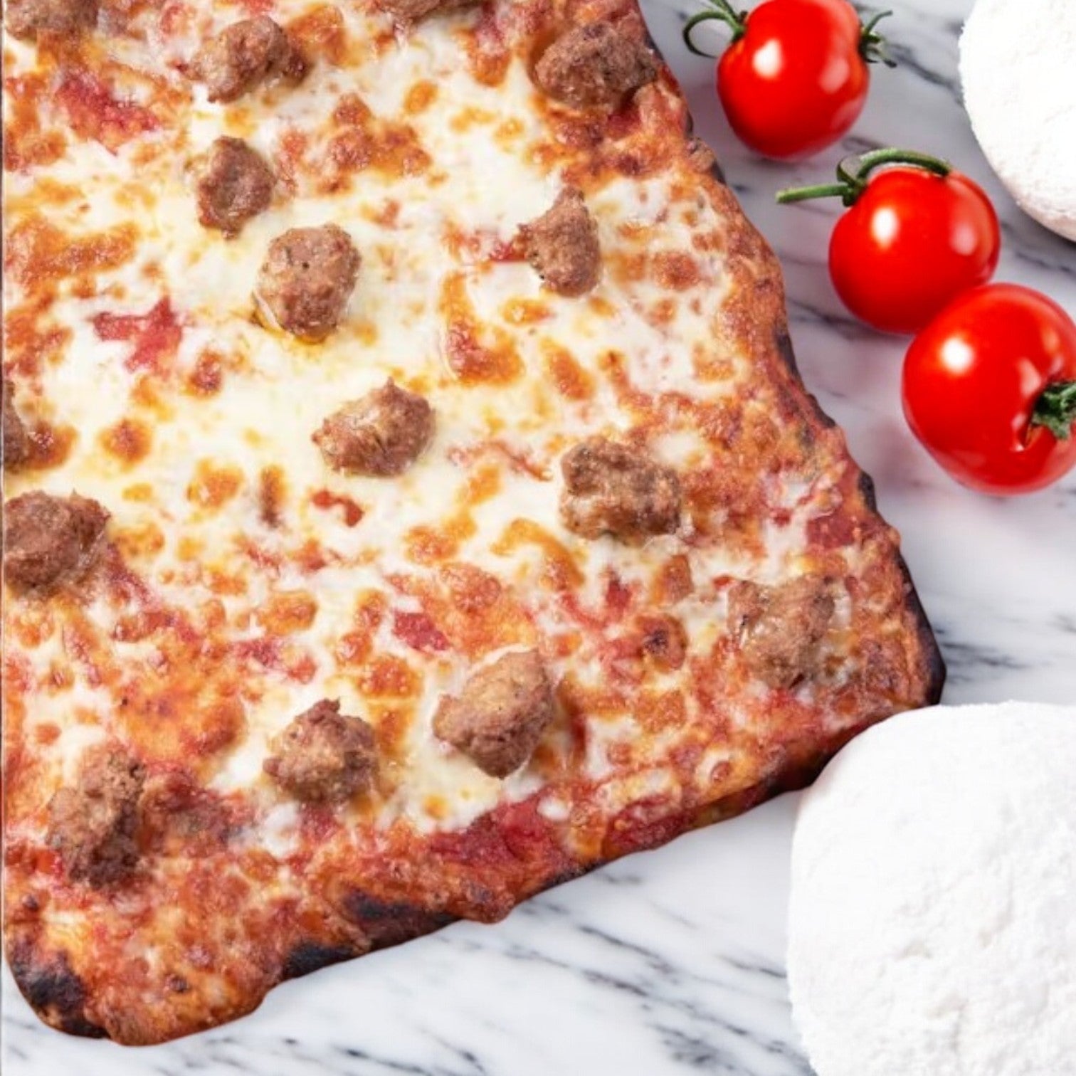 A rectangular Italian Sausage Pizza by Massa Pizza Co. sits on a marble surface, garnished with meatballs and cheese, alongside ripe red tomatoes and two balls of fresh dough.