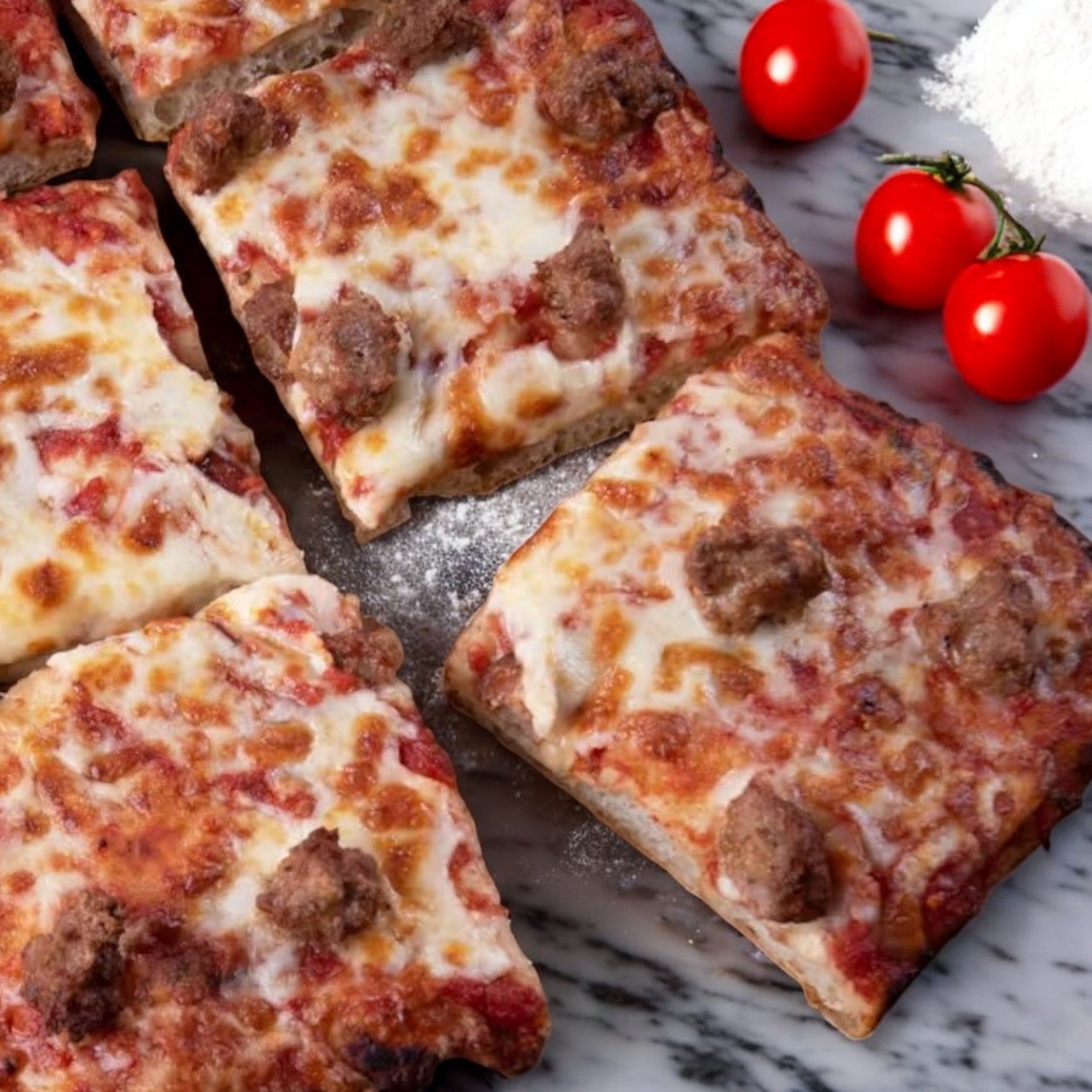 Square slices of Massa Pizza Co. Italian Sausage Pizza, a 21.6 oz. product from Massa Pizza, with cheese and sausage rest on a marble surface, embodying an authentic Italian flair. Next to them, cherry tomatoes glisten amid a sprinkle of flour, capturing the essence of bakery-style charm.