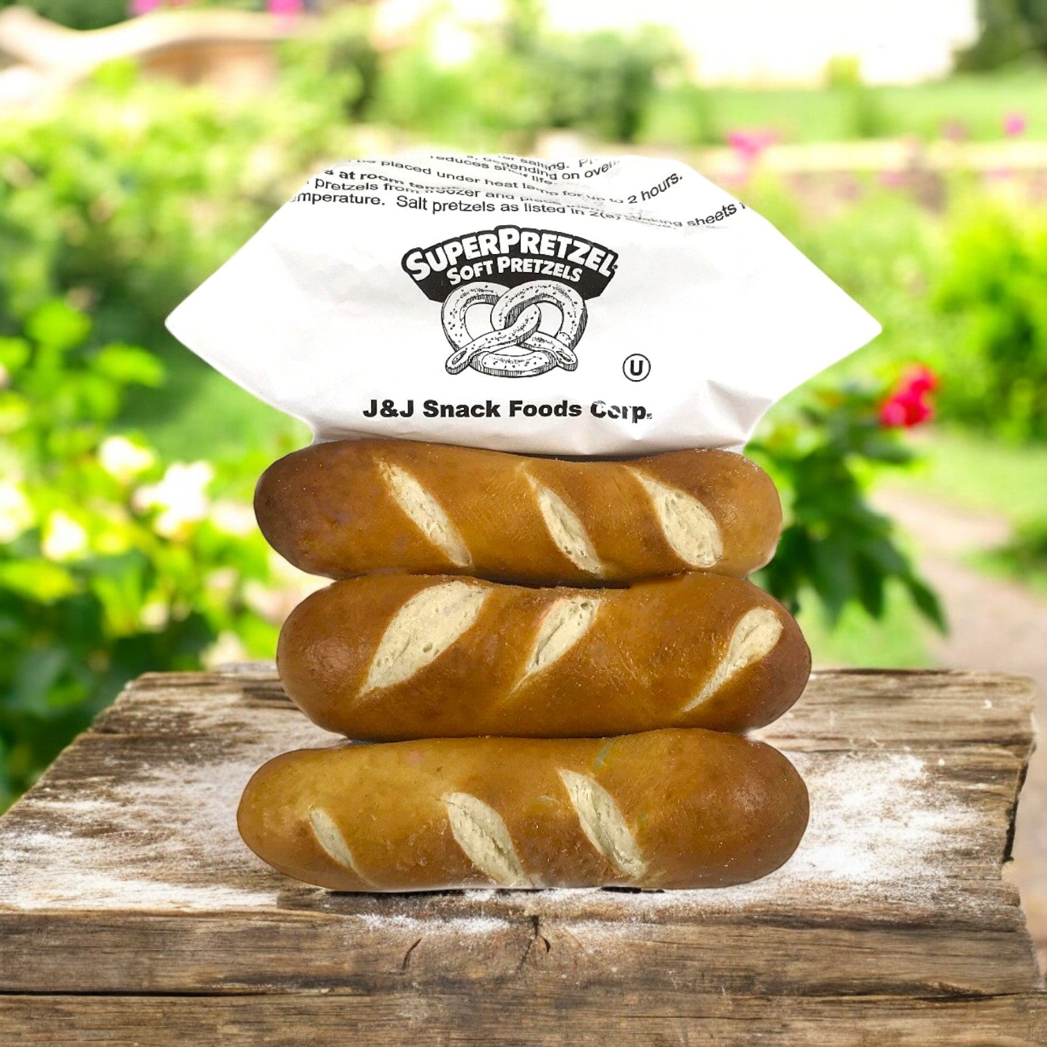 Three J&J Snacks Pretzel Bavarian Stick Minis stacked and wrapped in white paper sit on a wooden surface with a blurred garden backdrop—a perfect treat for satisfying snack cravings.