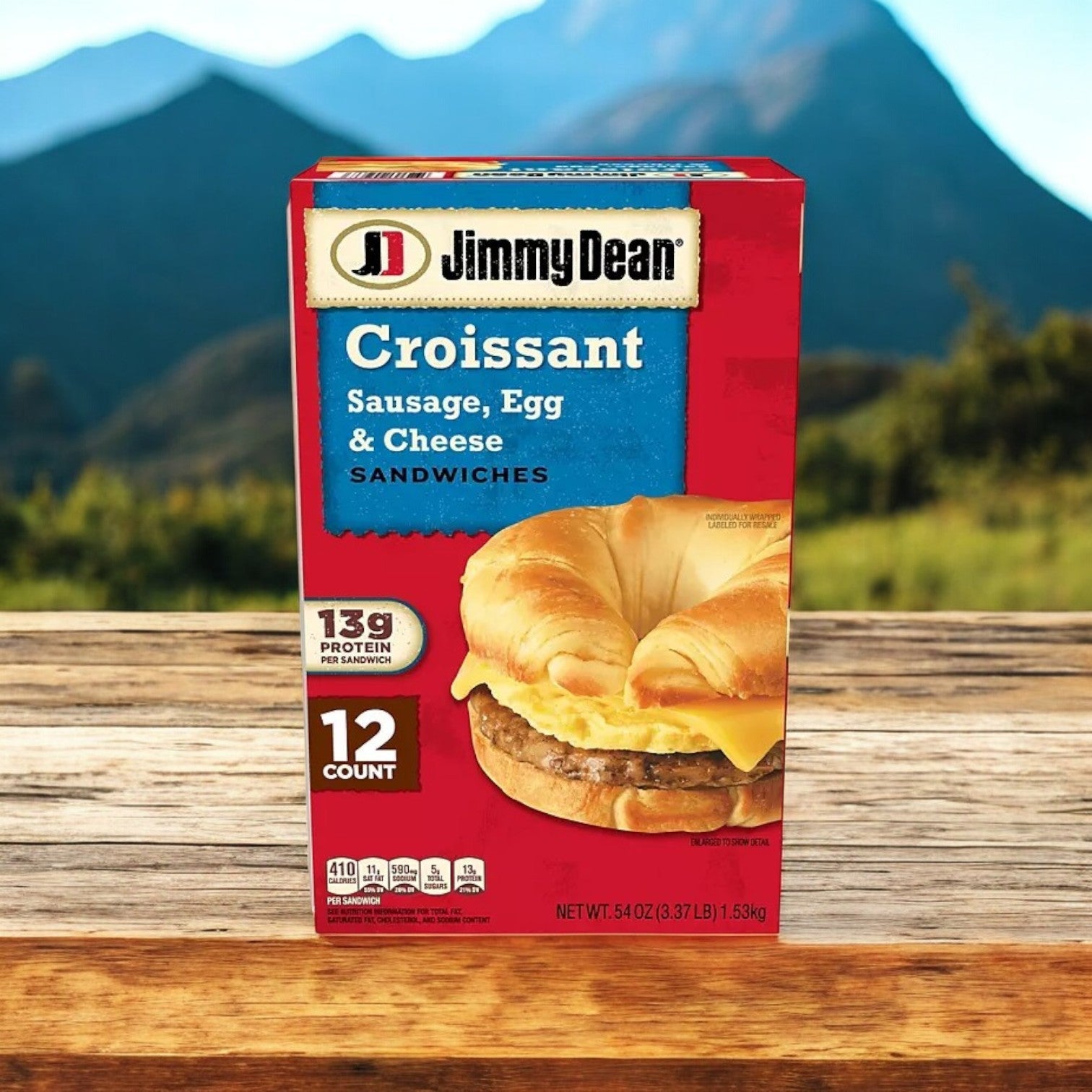 A box of Jimmy Dean Sausage, Egg & Cheese Croissant Sandwiches (12 ct) rests on a wooden surface with a stunning mountain backdrop, offering 12 protein-rich sandwiches with 13g of protein each for a satisfying start to your day.