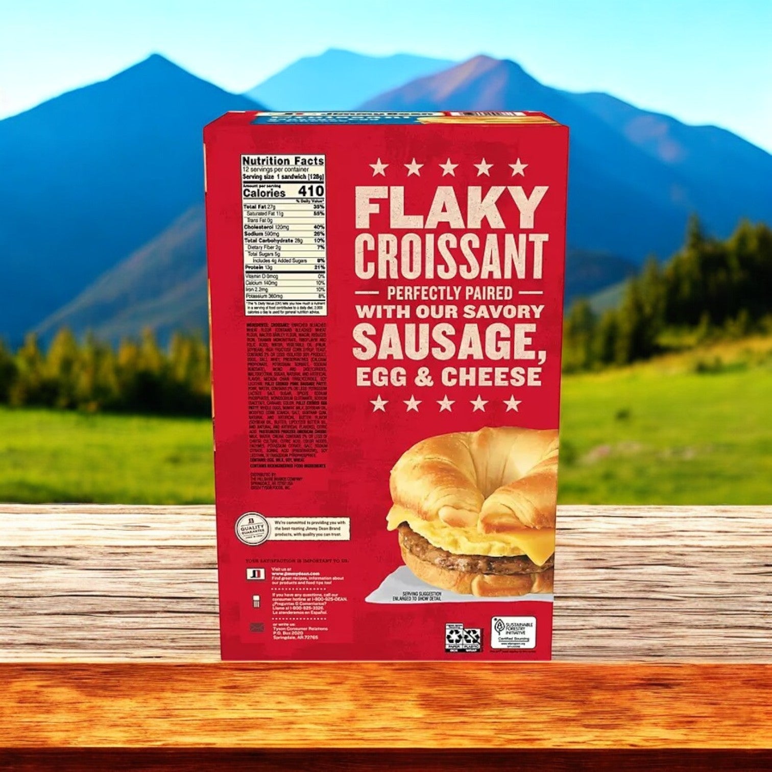A box of Jimmy Dean Sausage, Egg & Cheese Croissant Sandwiches (12 ct) rests on a wooden surface with majestic mountains behind, providing a quick and easy protein-packed breakfast for your busy mornings.