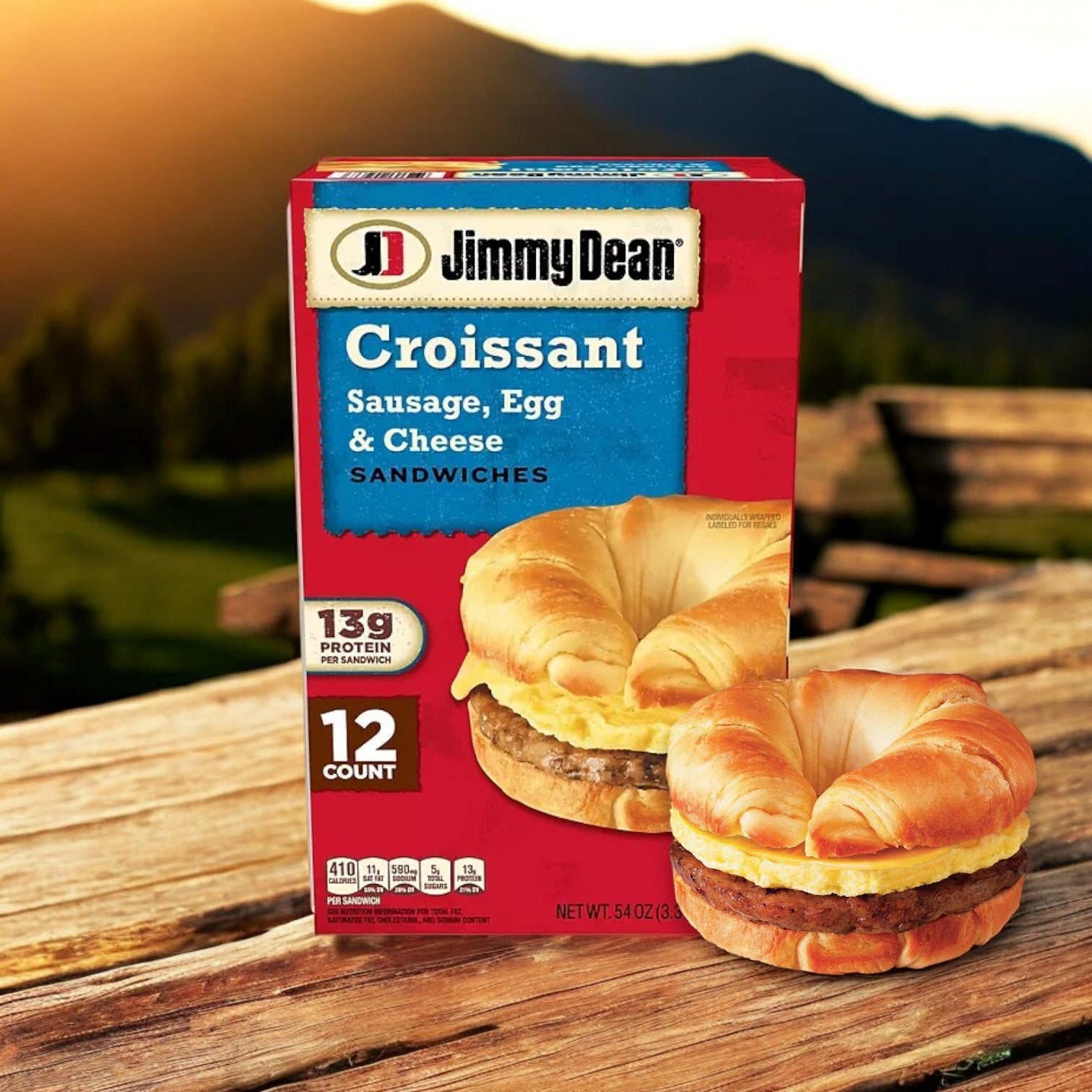 A box of Jimmy Dean Sausage, Egg & Cheese Croissant Sandwiches (12 ct) sits on a wooden table with one hearty sandwich beside it. In the background, there's blurred outdoor scenery of grass and mountains—a perfect quick and easy breakfast option for busy individuals.
