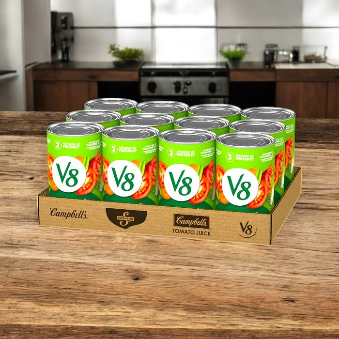 A wooden kitchen countertop showcases a cardboard tray containing 12 cans from the "V8" brand's 46 oz. Vegetable Juice, wrapped in green labels. Savor each can's heart-healthy benefits of 100% vegetable juice.