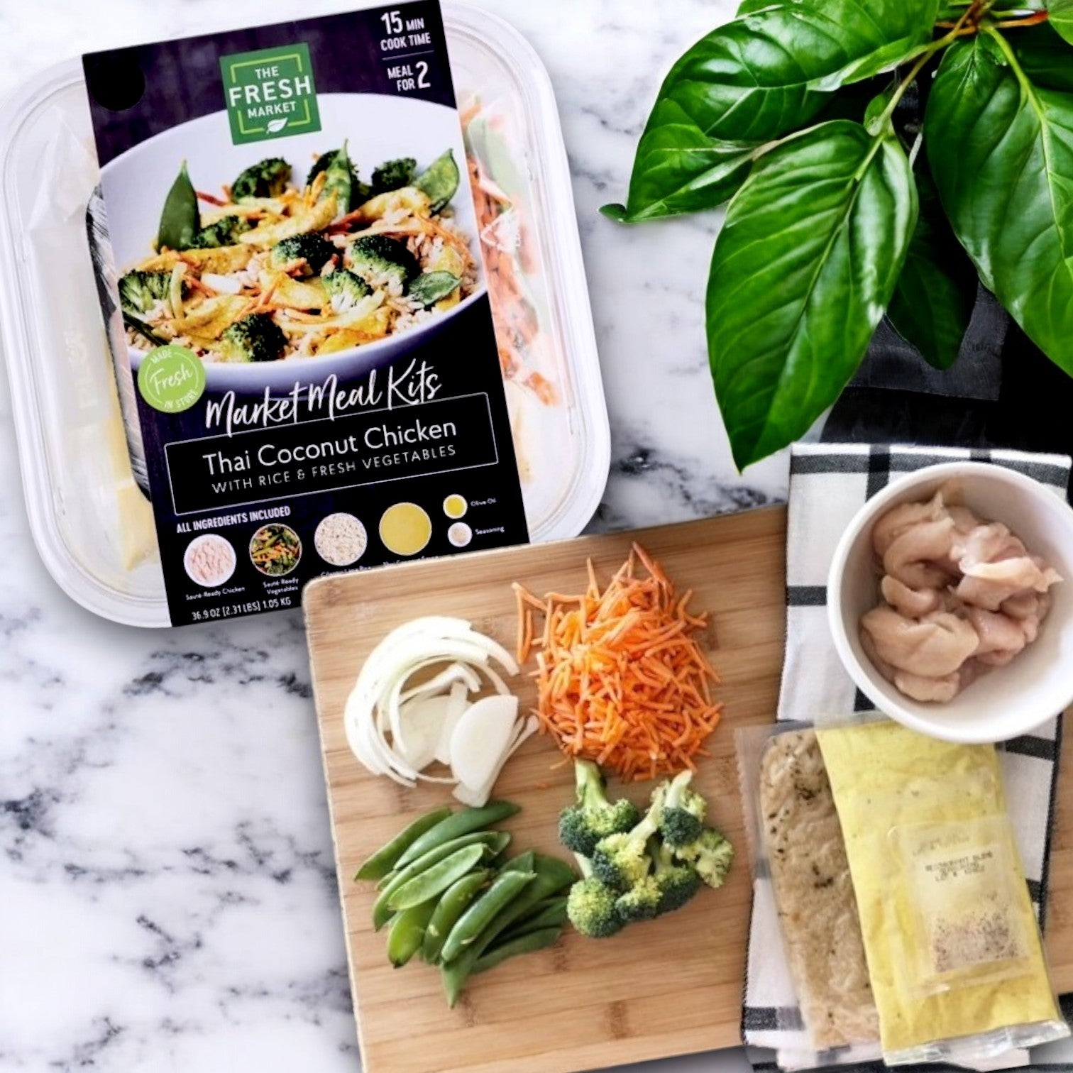 The Fresh Market Thai Coconut Chicken with Rice & Fresh Vegetables Market Meal Kit is presented on a marble surface, featuring packaged ingredients and sliced vegetables for authentic Thai flavors. A green plant adds a fresh touch to this culinary experience.