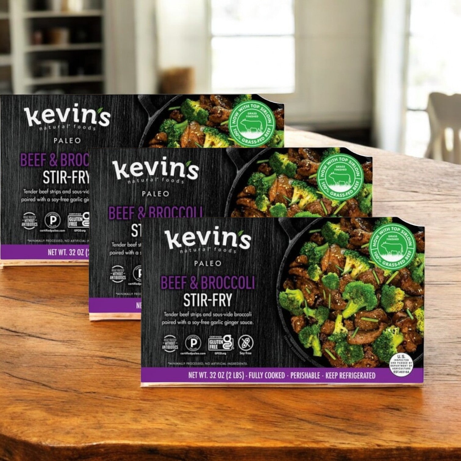 Three packs of Kevin's Beef & Broccoli Stir-Fry (32 oz - 3 Count) sit on a wooden surface, displaying the label and enticing food image. Crafted with quality ingredients, this meal is perfect for busy days.