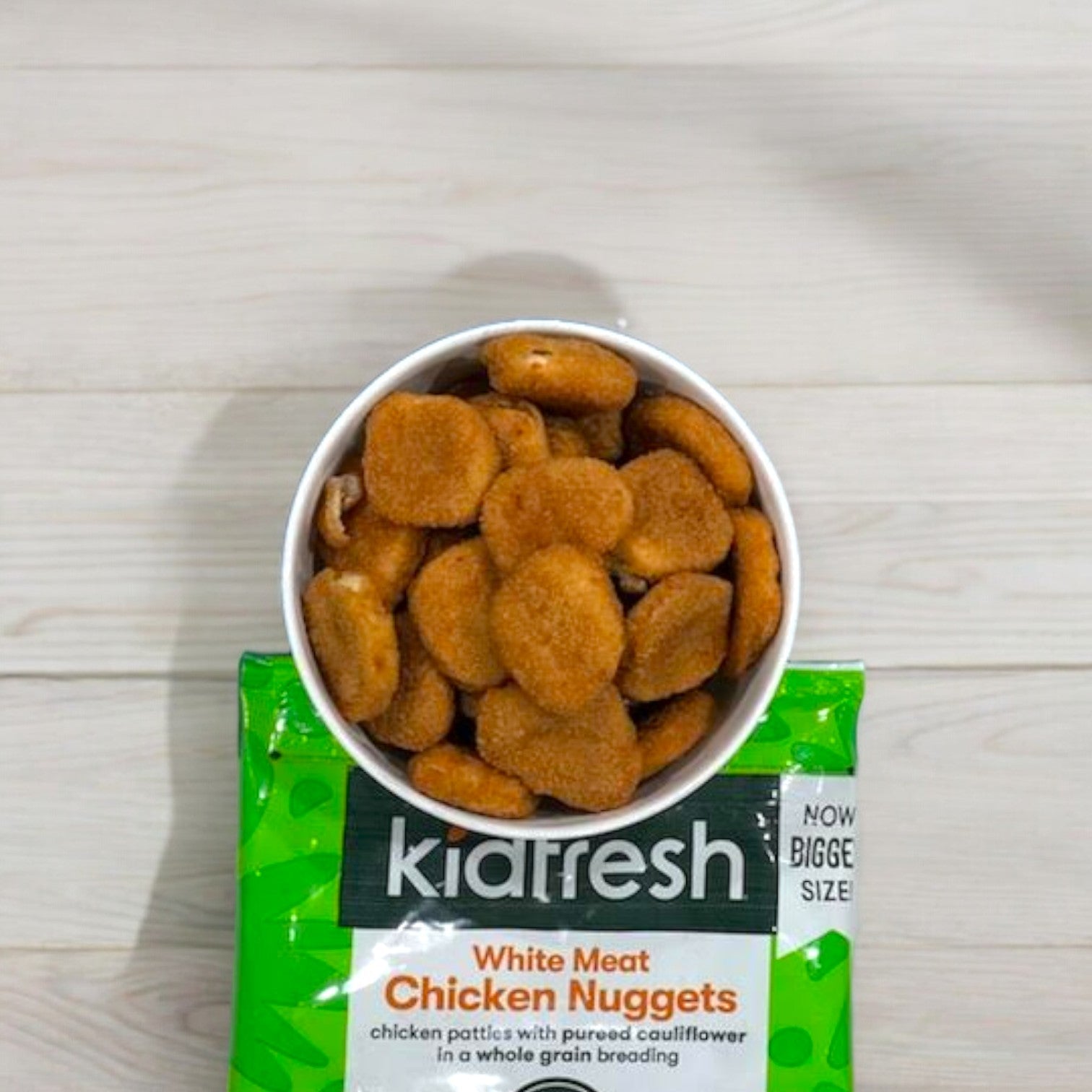 A bowl filled with Kidfresh Frozen Chicken Nuggets - 16.35 oz sits on a green Kidfresh packaging labeled "White Meat Chicken Nuggets." The package highlights "Now Bigger Size" and mentions hidden veggies like cauliflower in the breading.