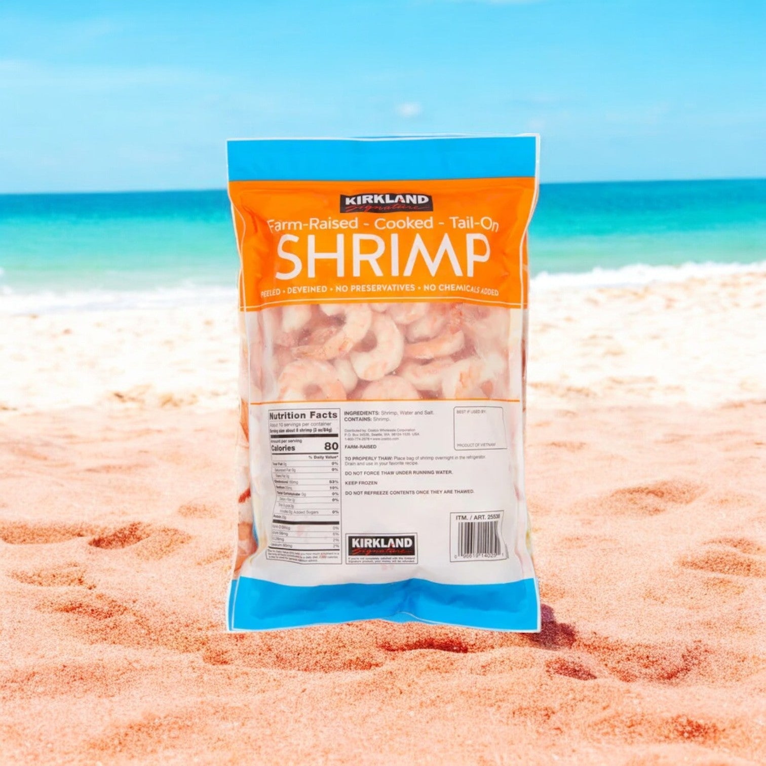 A package of Kirkland Signature Farm-Raised Cooked Shrimp is positioned upright on a sandy beach, with the ocean and blue sky in the background, highlighting its premium quality and farm-raised origins.