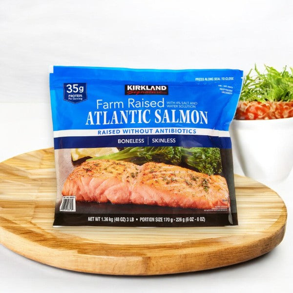 A 3 lb pack of Kirkland Signature Atlantic Salmon (6 oz - 8 oz center-cut portions) sits on a wooden tray with greens nearby, emphasizing its omega-3 richness.