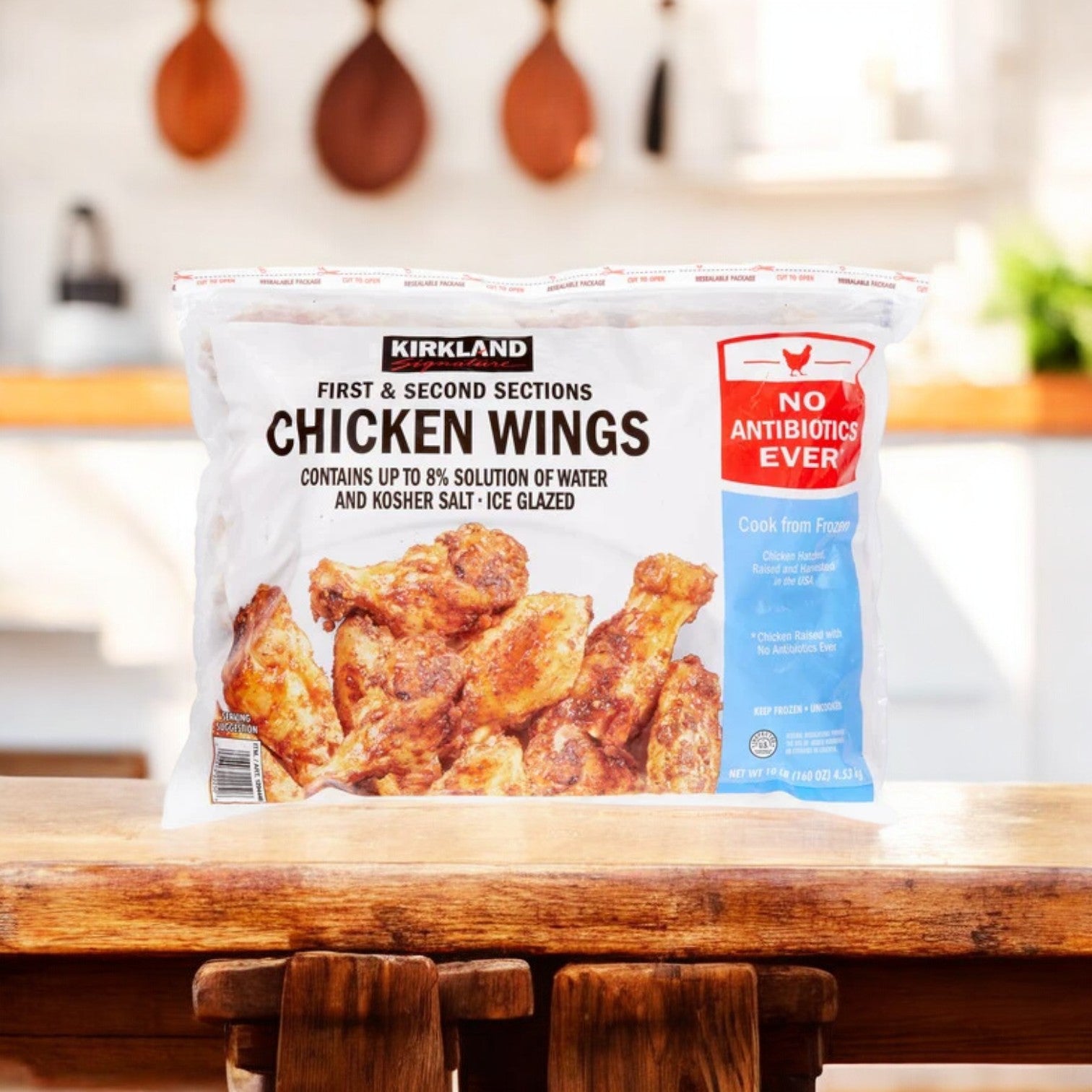 A 10-lb Kirkland Signature bag of frozen chicken wings labled "No Antibiotics Ever" and "First and Second Sections" rests on the kitchen table. These quality cuts provide versatile cooking options for any meal.