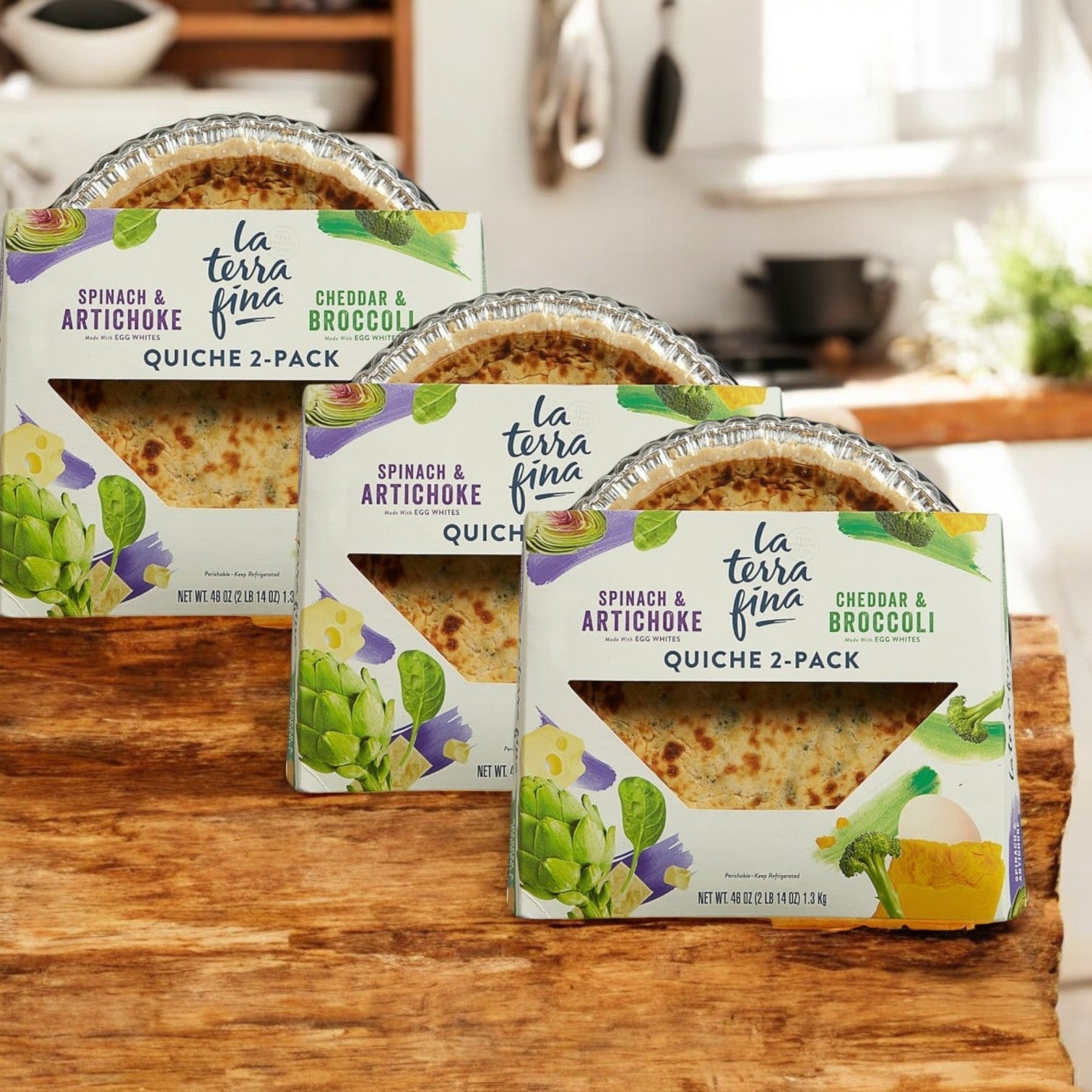 Three boxes of La Terra Fina Quiche, 46 oz. each, in Spinach & Artichoke and Cheddar & Broccoli flavors, are on a wooden kitchen table.