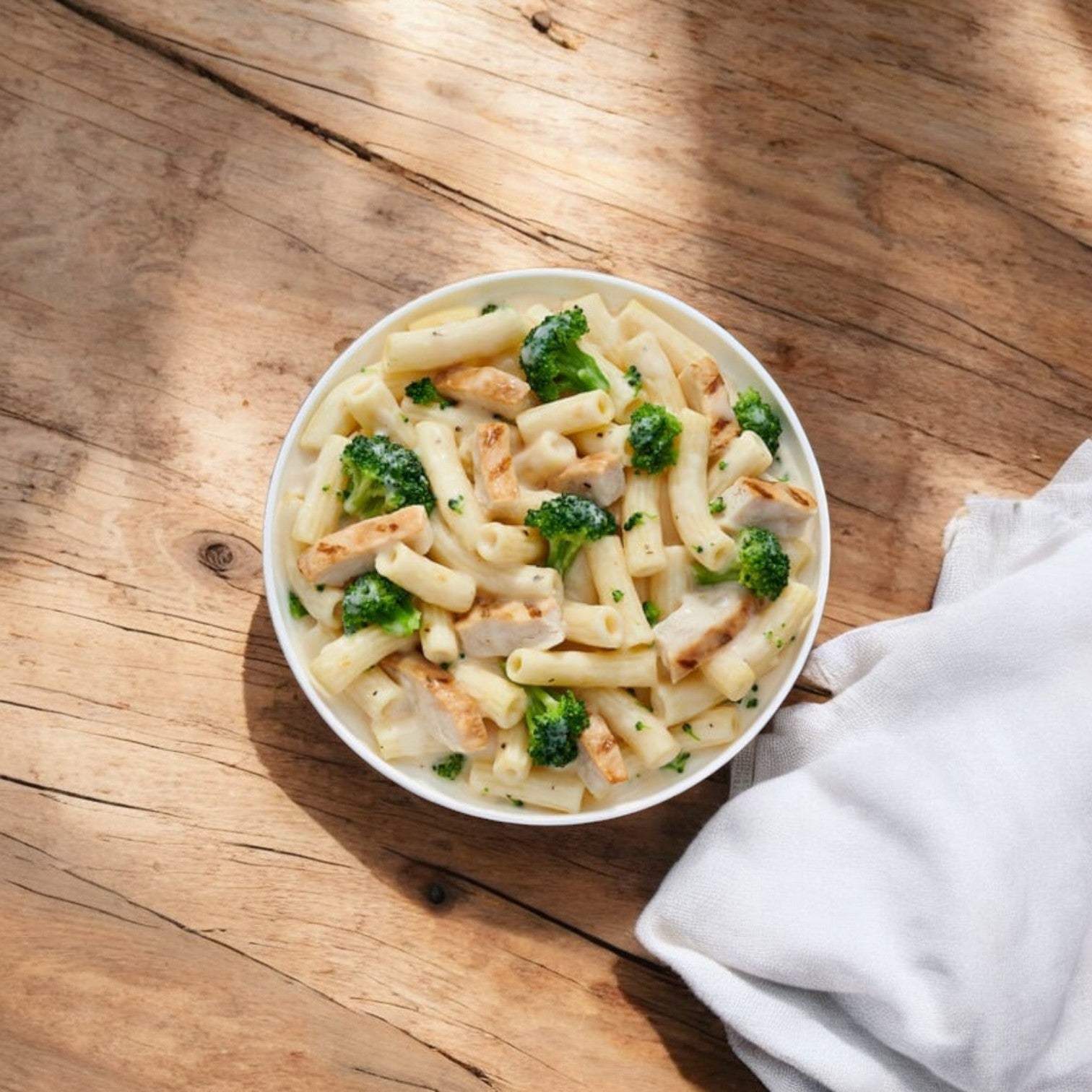 A bowl of Lean Cuisine Alfredo Pasta with Chicken & Broccoli on a wooden surface next to a white cloth, resembling a delightful microwaveable entrée.