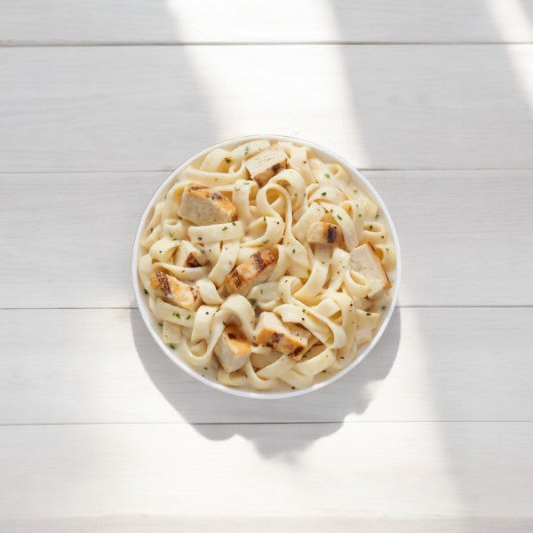 A white bowl filled with fettuccine Alfredo topped with grilled chicken pieces lies on a white wooden surface, akin to a serving of Lean Cuisine Favorites Chicken Fettuccini.