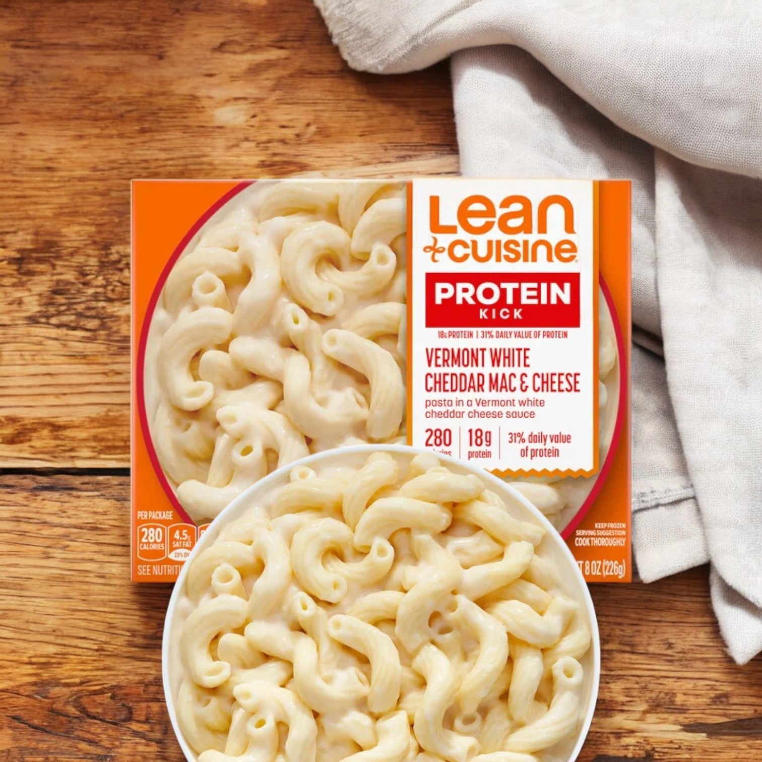 Photograph of Lean Cuisine Features Vermont White Cheddar Mac And Cheese meal tray on a wooden surface. The packaging emphasizes its protein content, calorie count, and distinct white cheddar flavor. This creamy pasta dish is an 8 oz. microwaveable meal, perfect for quick and delicious dining.