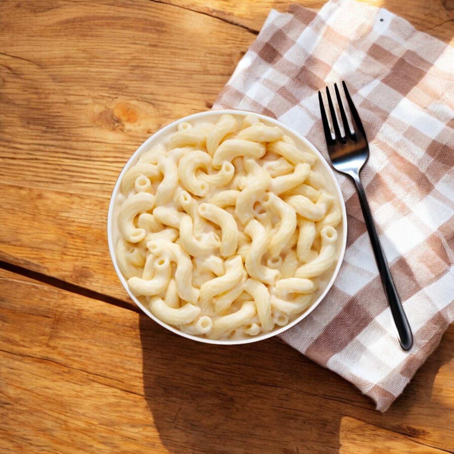 A bowl of Lean Cuisine’s Vermont White Cheddar Mac And Cheese is placed on a checkered cloth with a fork beside it on a wooden table.