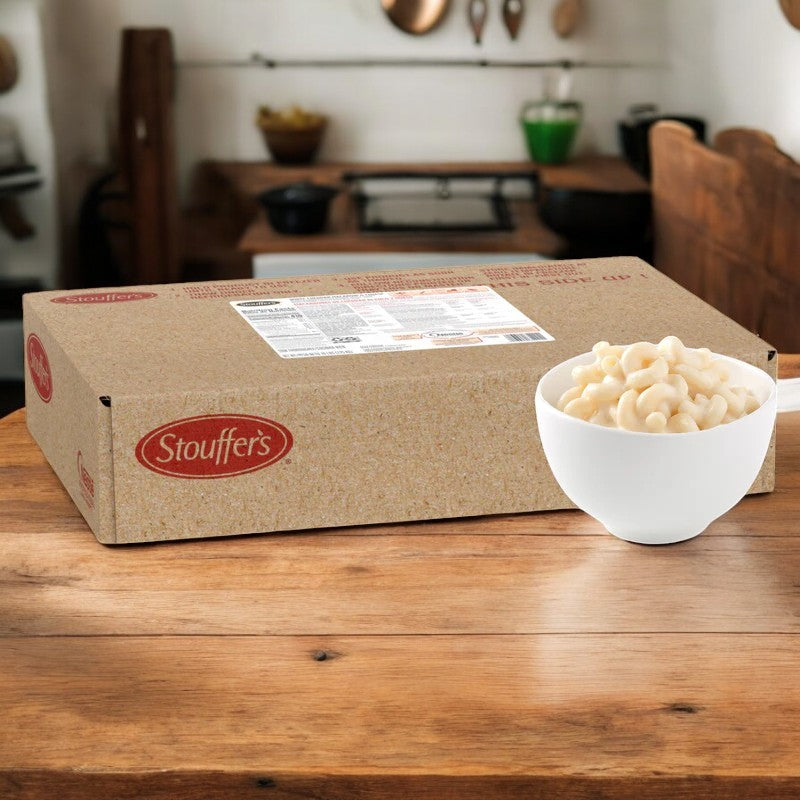A case of Stouffer's Macaroni and Cheese White, 64 oz., rests on a wooden table alongside a white bowl filled with cheesy delight. The kitchen backdrop enhances the warmth of this shareable family-sized meal.