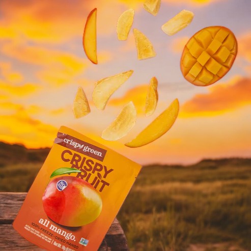 A packet of Crispy Green Crispy Fruit All Mango - 0.35 oz - 1 Count on a table with dried mango slices and a fresh mango above, set against a sunset background, showcases an array of sustainable snacks.