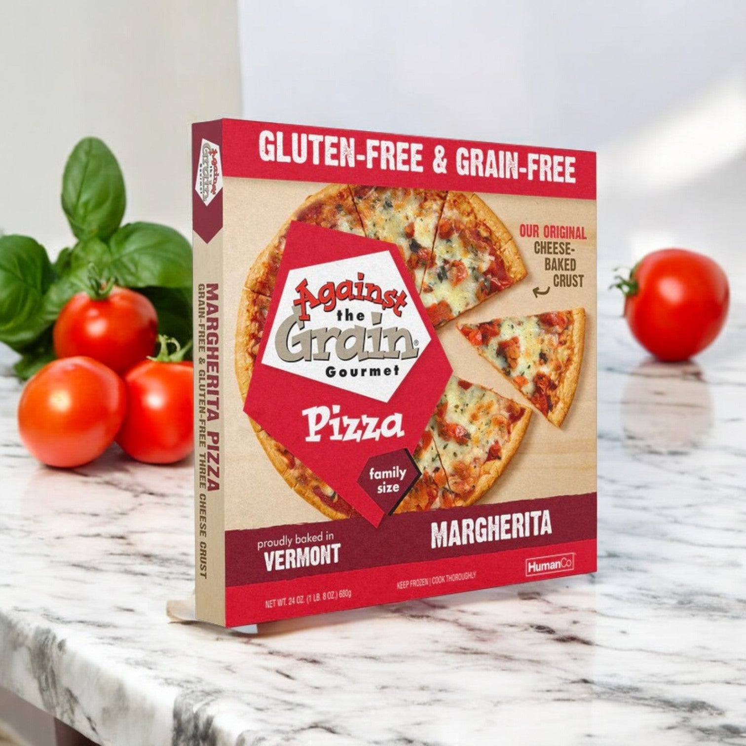 On a marble surface, the "Against the Grain Gourmet Pizza, Margherita" box is surrounded by fresh tomatoes and basil. This 24 oz gourmet pizza is gluten-free and grain-free, offering a family-sized option for a delightful culinary experience.
