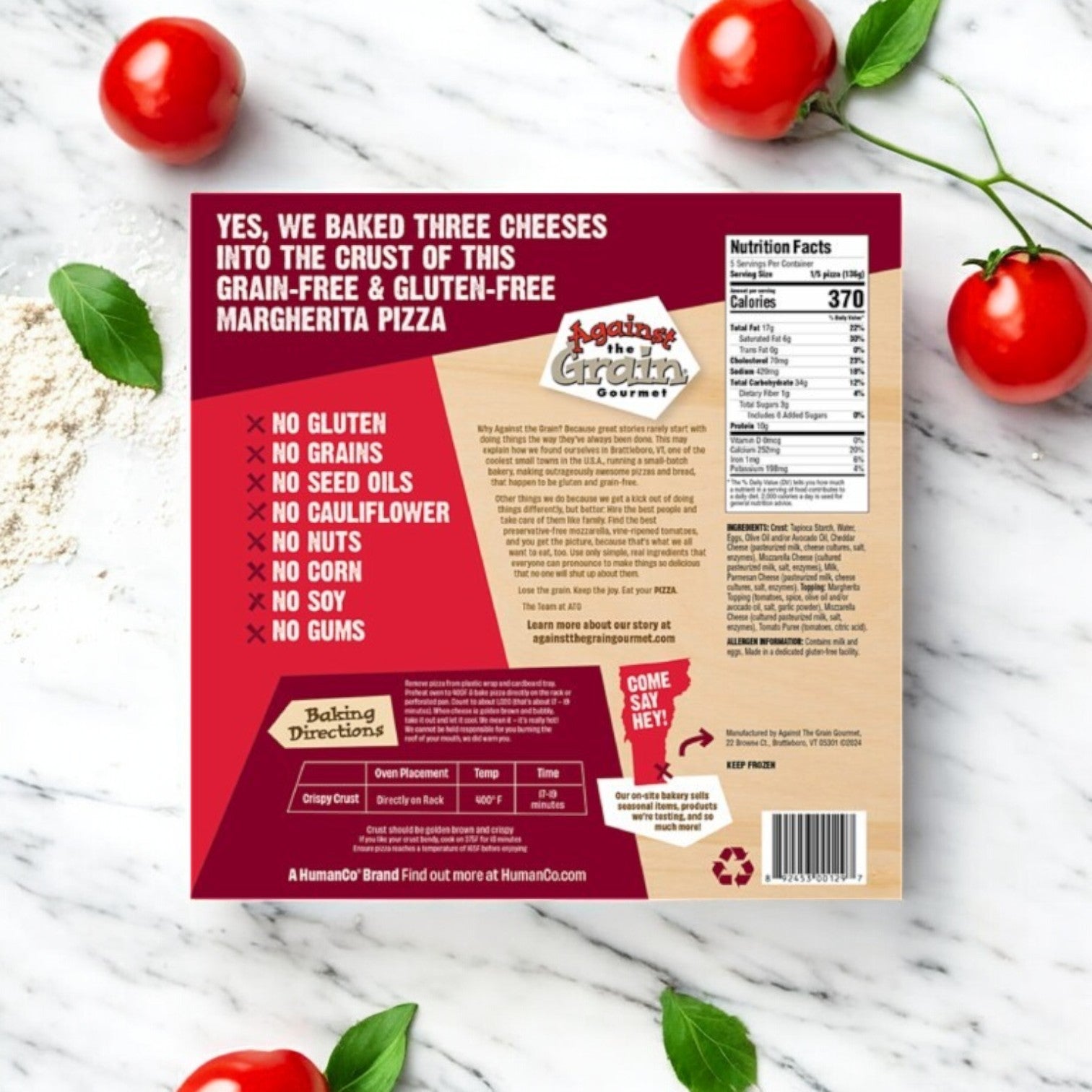 The back of the Against the Grain Gourmet Margherita Pizza box displays nutrition facts, a gluten-free ingredient list, and baking directions. It is elegantly surrounded by fresh tomatoes, aromatic basil leaves, and a dusting of flour on a pristine white surface.